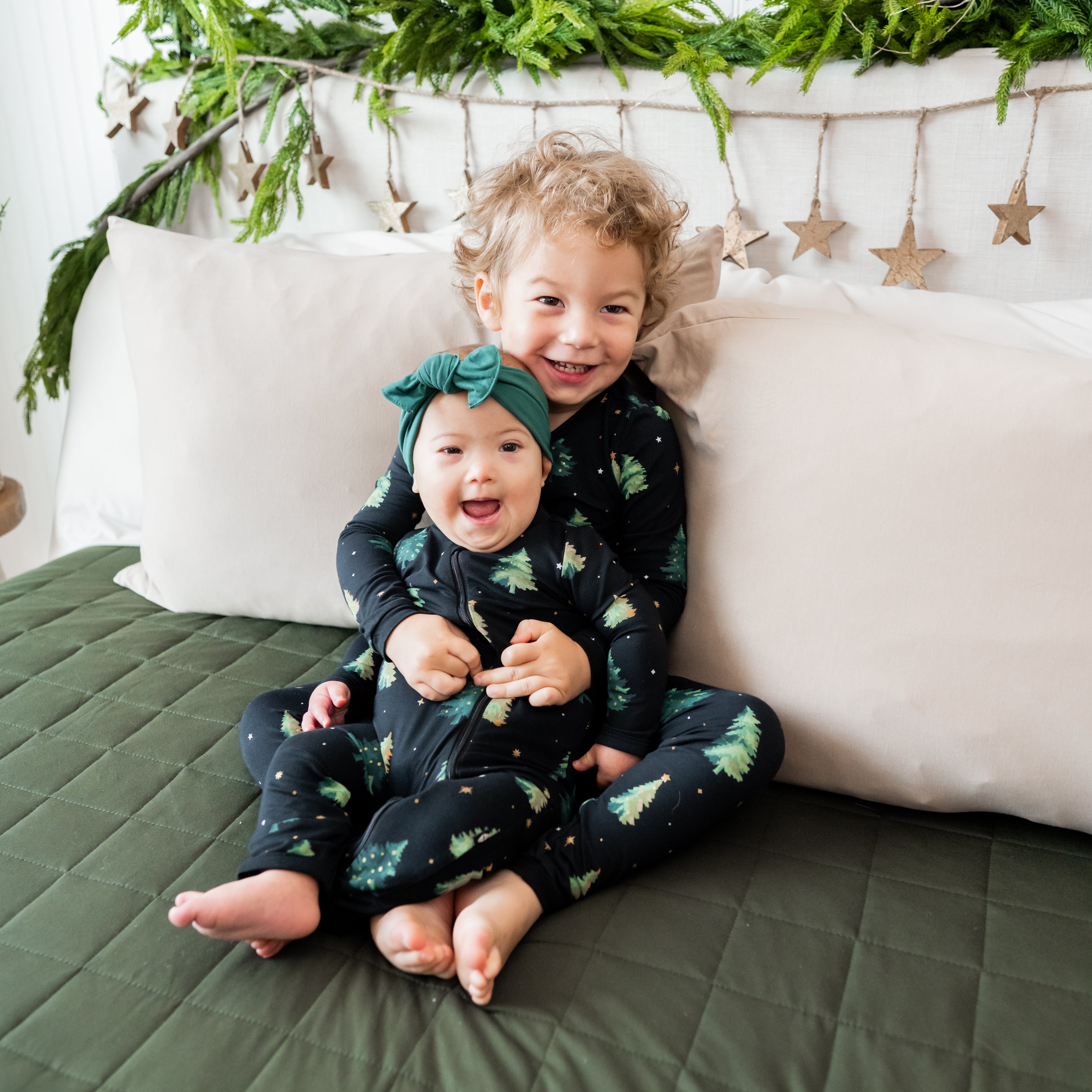 Brother holding sister both sitting on a bed wearing matching pajama set and Zippered Romper in Twinkle Tree