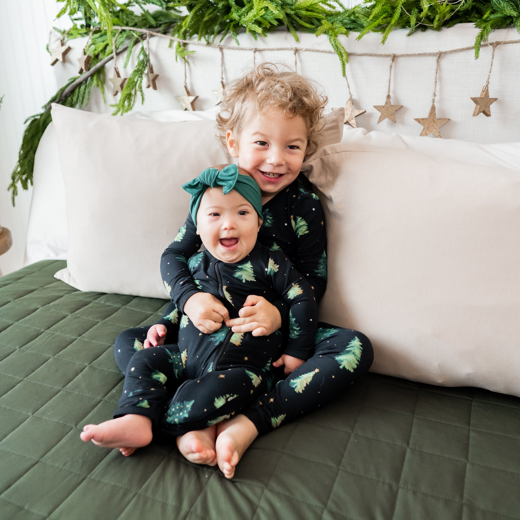Brother holding sister both sitting on a bed wearing matching pajama set and Zippered Romper in Twinkle Tree