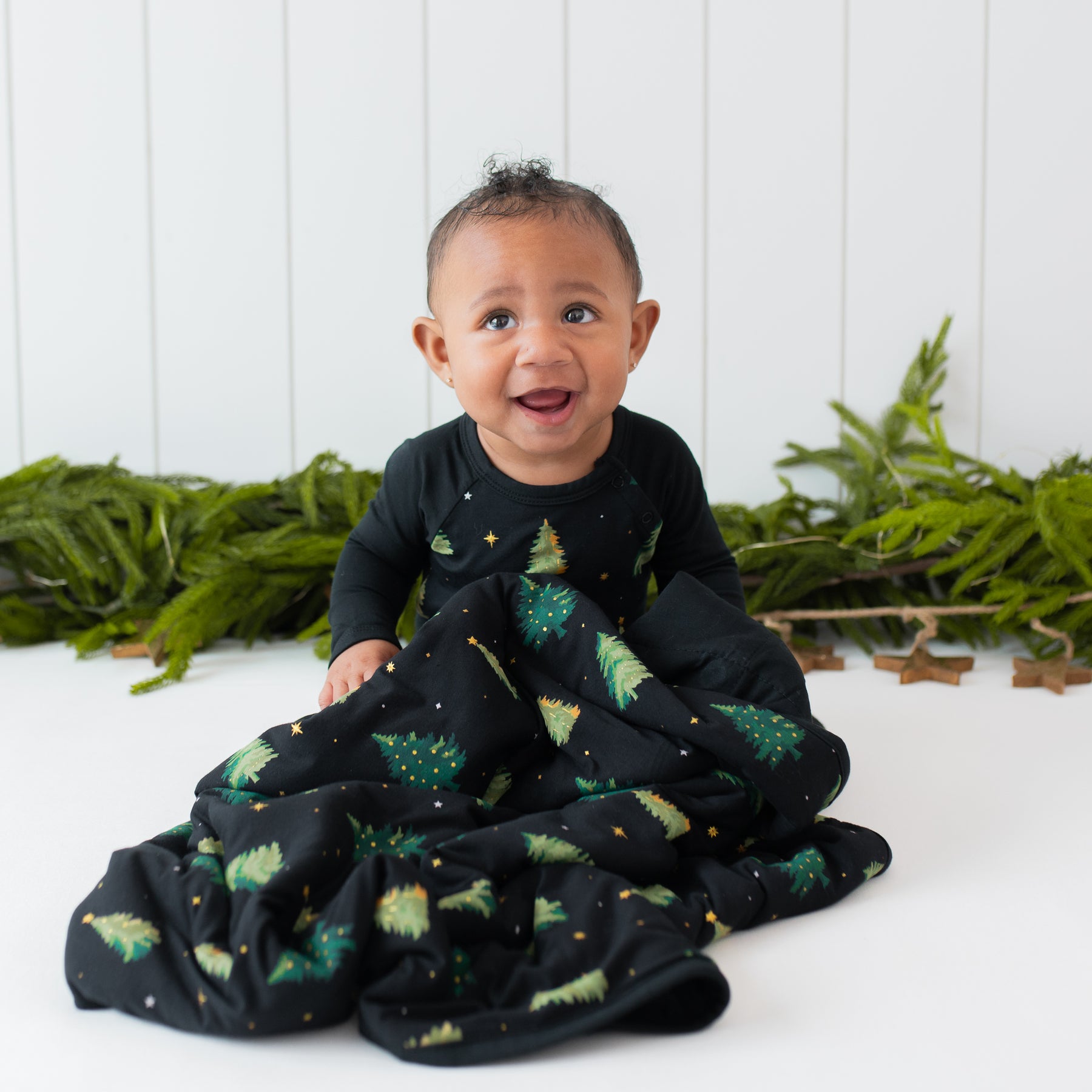 Infant sitting in front of greenery with a Baby Blanket in Twinkle Tree wearing a matching long all