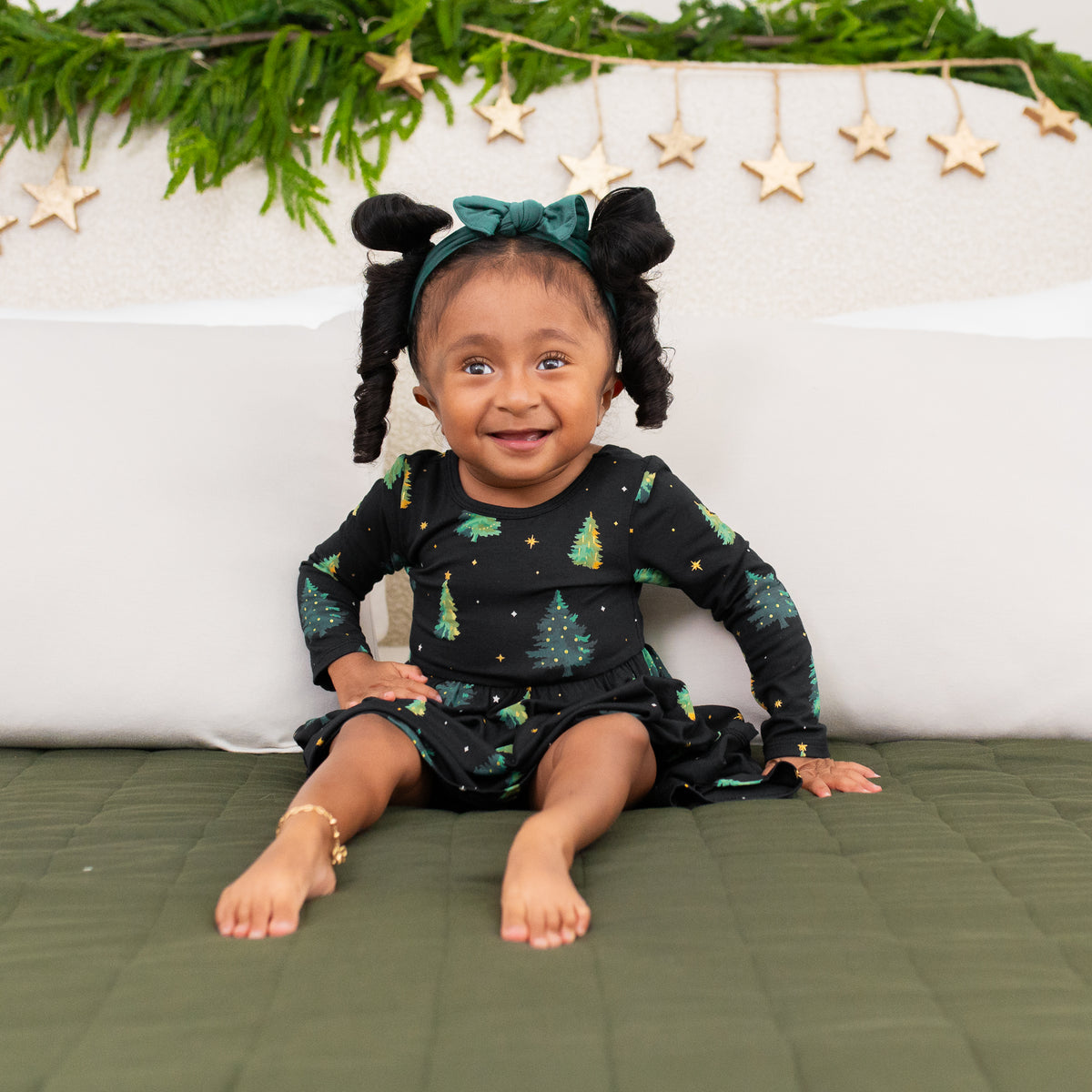 Young toddler sitting on a bed wearing the Long Sleeve Twirl Bodysuit Dress in Twinkle Tree and Emerald knotted bow headband
