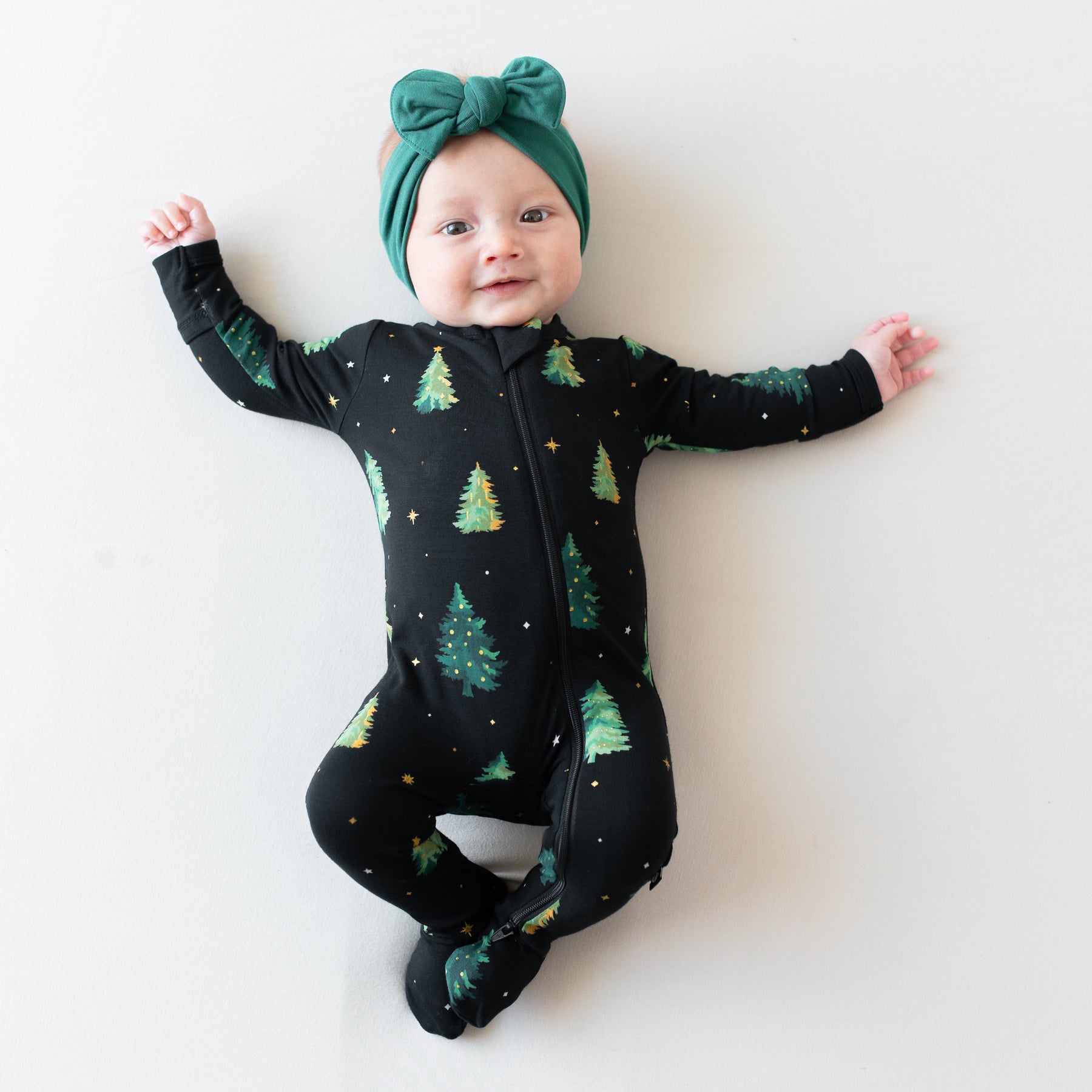 Infant laying down wearing the Zippered Footie in Twinkle Tree with Emerald knotted bow headband