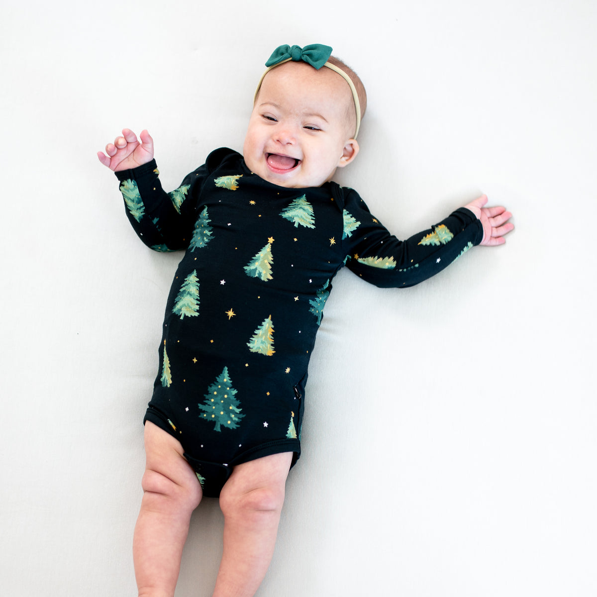 Infant lying down wearing the Long Sleeve Bodysuit in Twinkle Tree and tiny nylon bow in Emerald