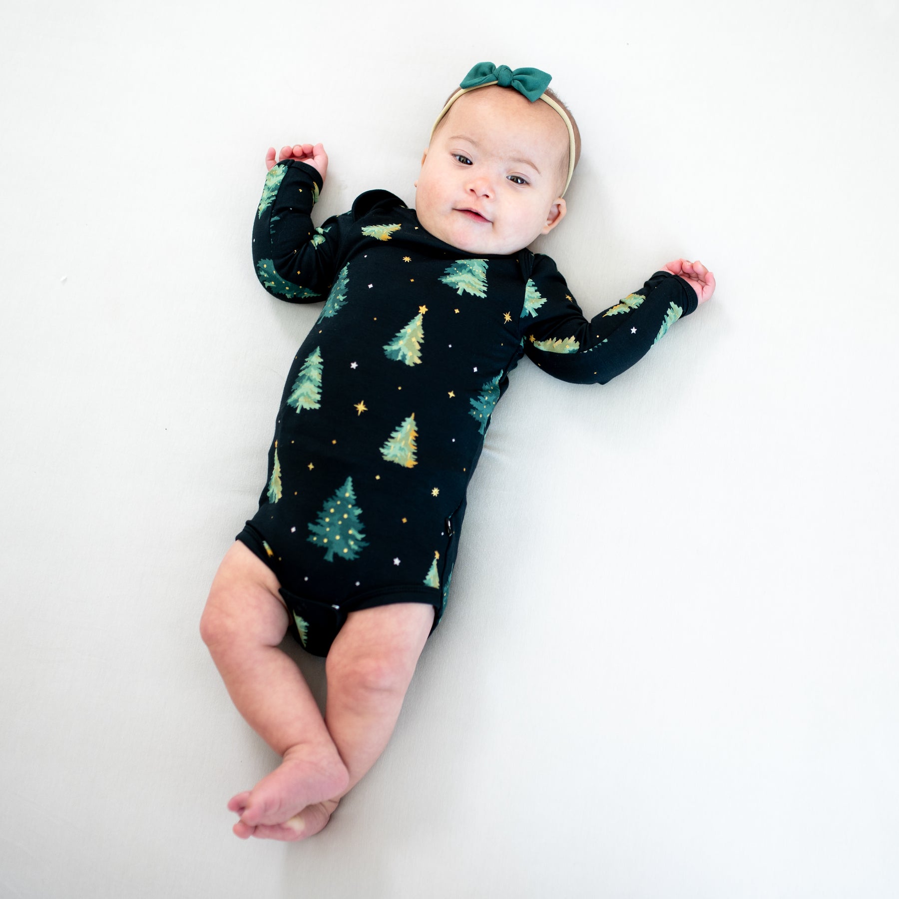 Infant lying down wearing the Long Sleeve Bodysuit in Twinkle Tree with a tiny nylon bow in Emerald