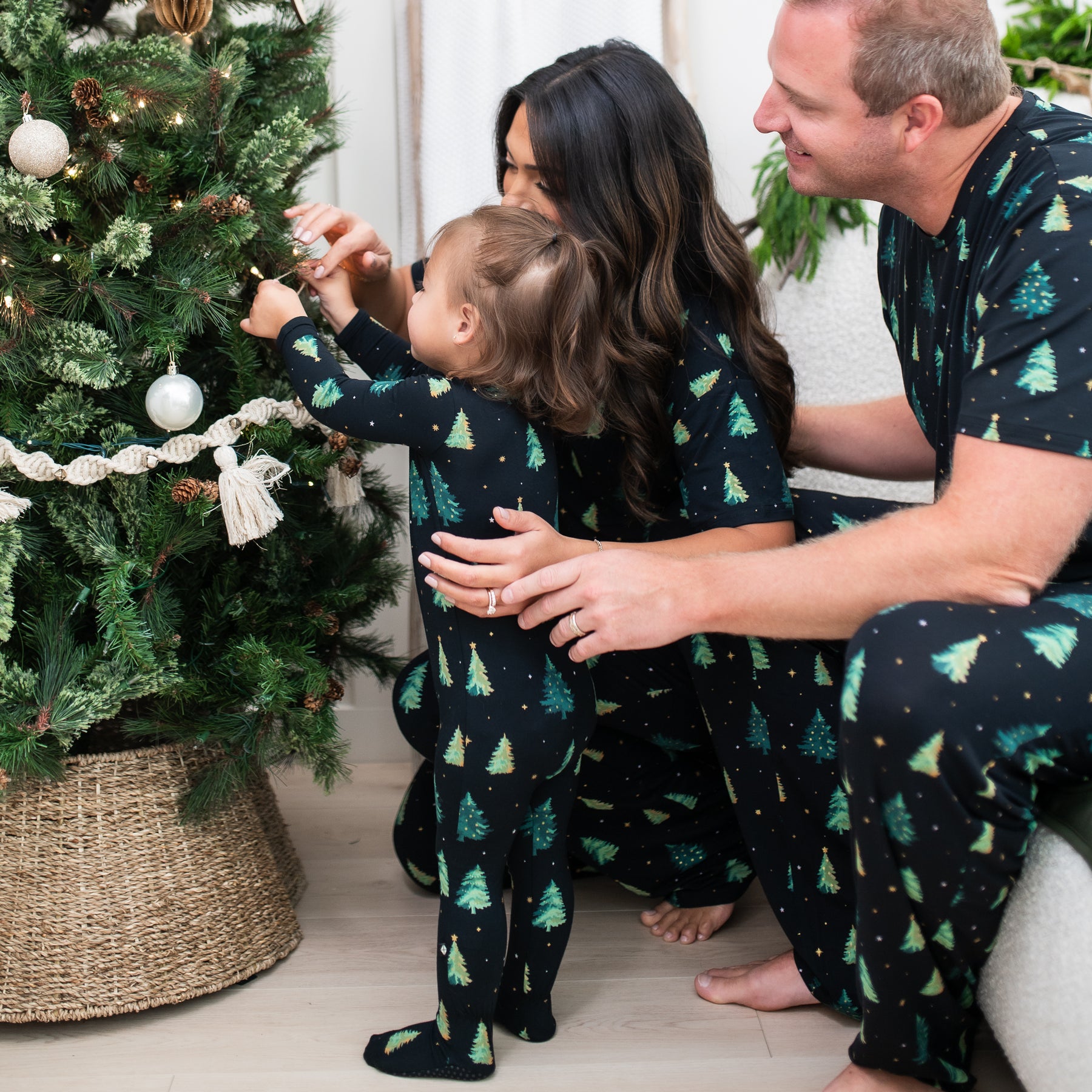Family of 3 matching in Twinkle Tree sitting around a Christmas tree while the parents watch their daughter put a christmas ornament on a christmas tree