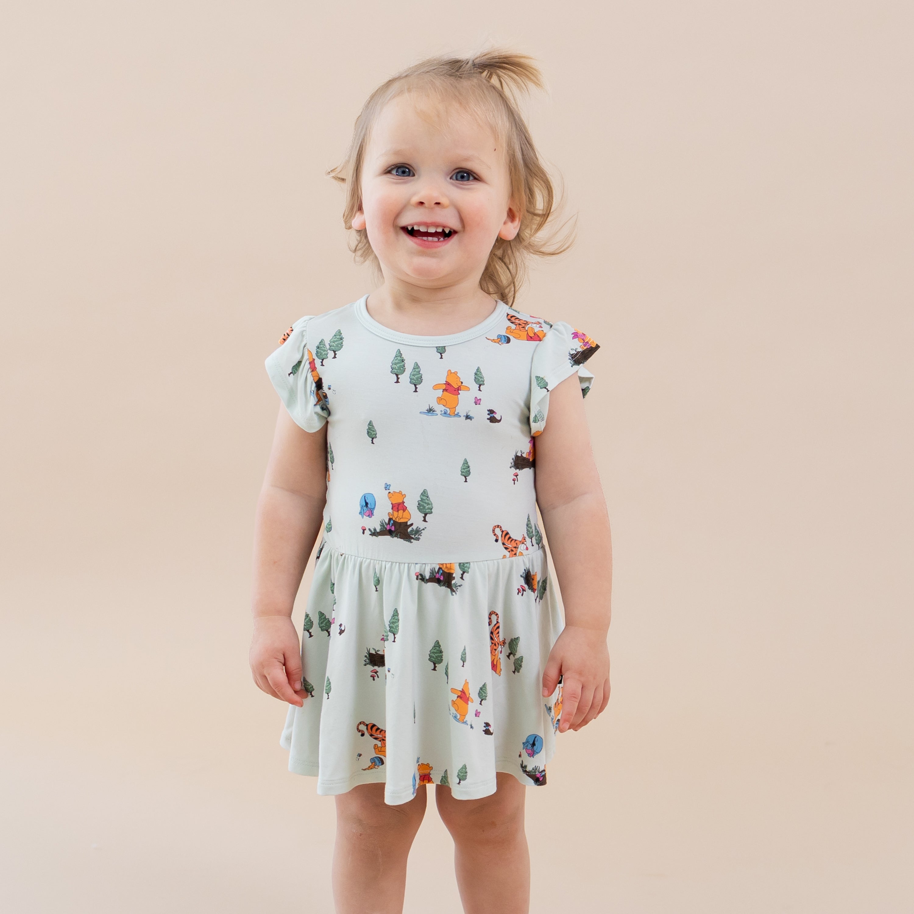Young toddler wearing the Twirl Bodysuit Dress in Winnie the Pooh Hundred Acre Wood standing in front of a light neutral surface