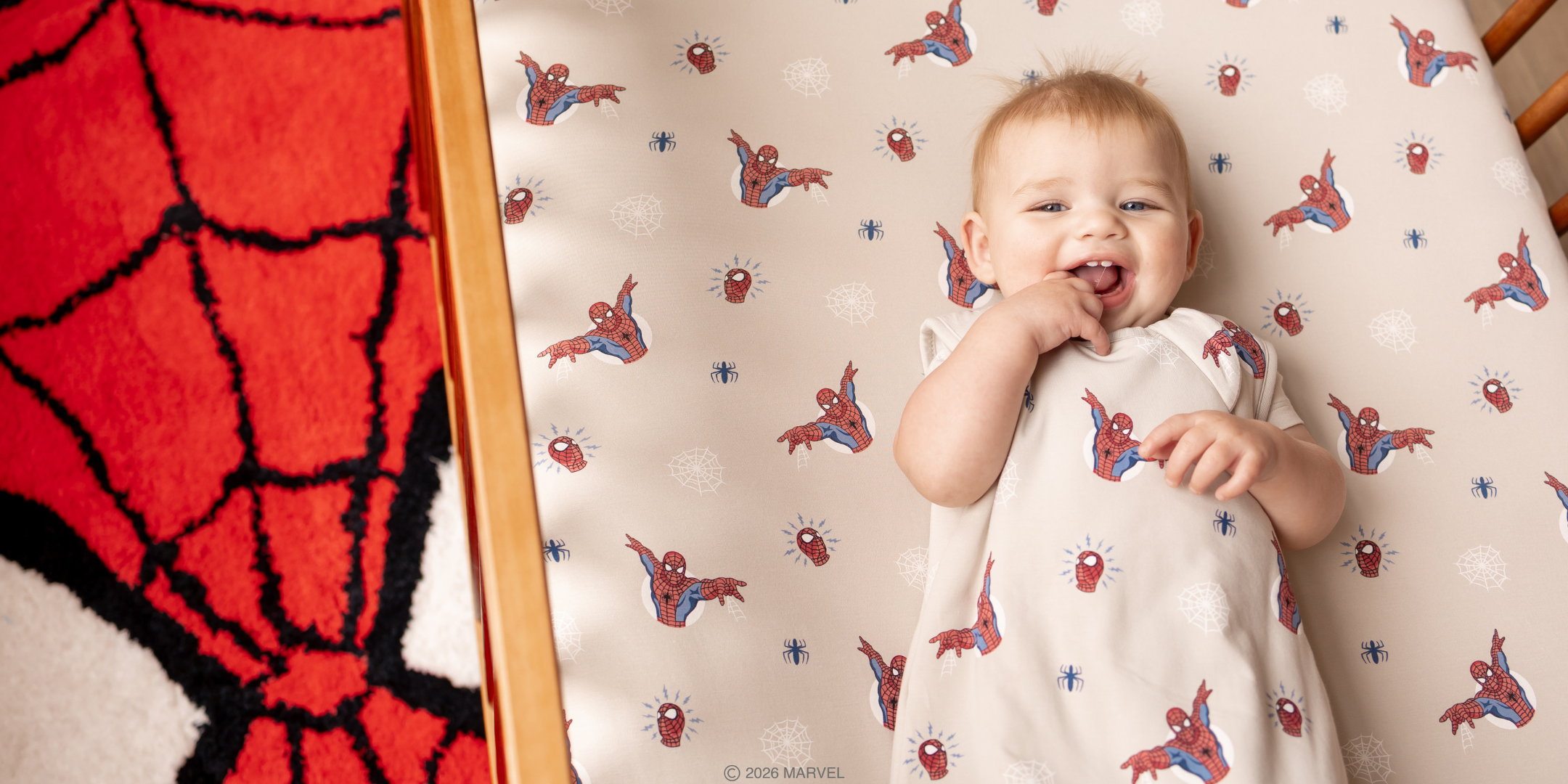 A baby in a crib wearing a Spiderm-Man inspired sleep sack with matching crib sheet on mattress and Spider-Man rug on the floor