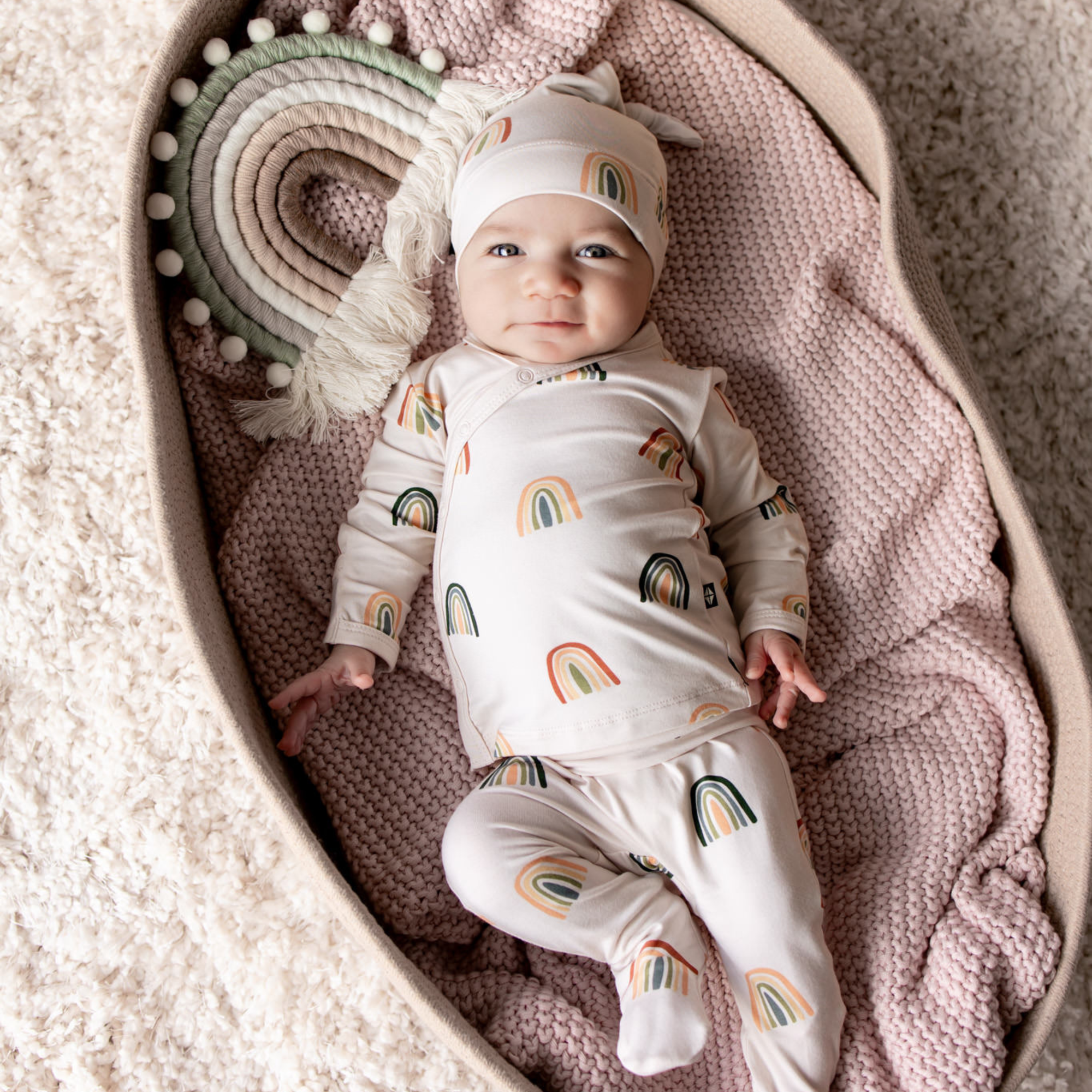 Baby in a take me home set with kimono top, pants and matching cap in a neutral rainbow pattern laying in a bassinet 