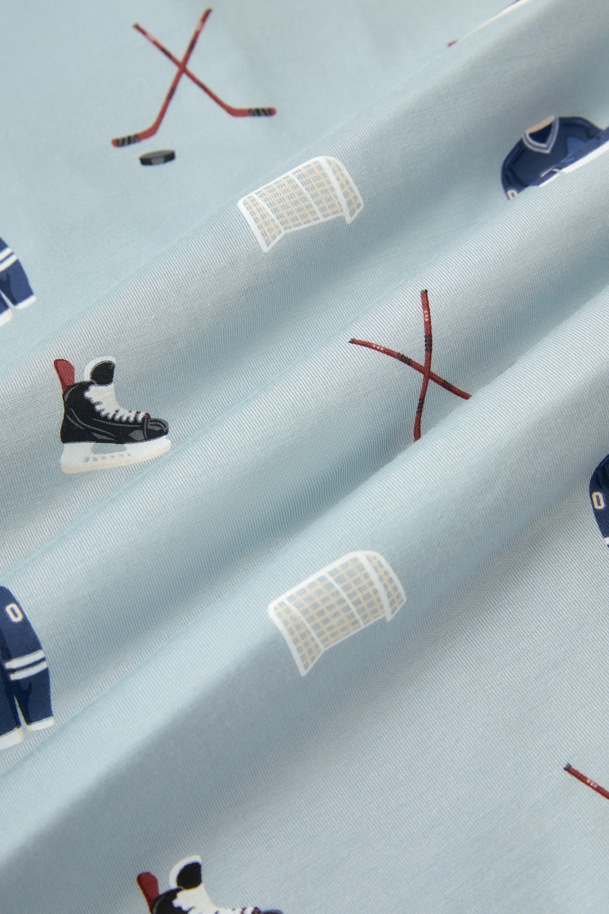 A repeating, all-over print featuring a dark blue hockey jersey with the numbers 00, a pair of crossed brown hockey sticks with a black hockey puck, a white hockey goal, and a pair of black hockey skates, over a light dusty blue background