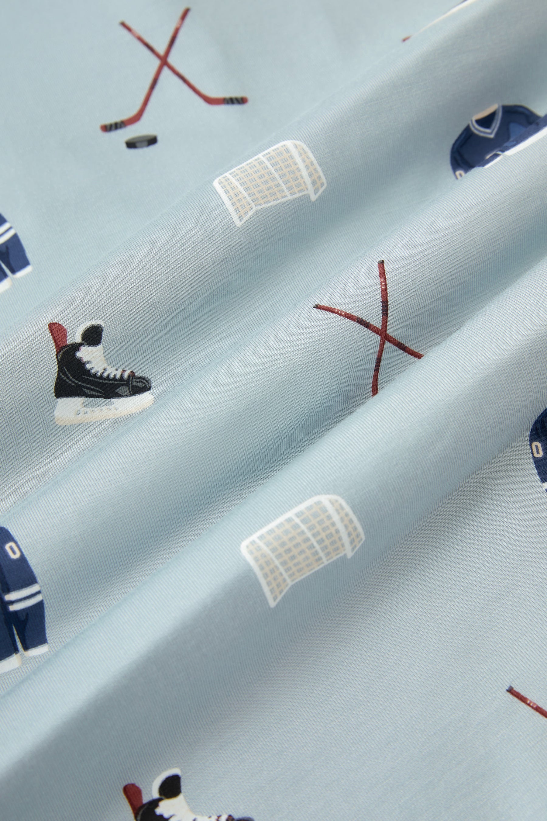 A repeating, all-over print featuring a dark blue hockey jersey with the numbers 00, a pair of crossed brown hockey sticks with a black hockey puck, a white hockey goal, and a pair of black hockey skates, over a light dusty blue background