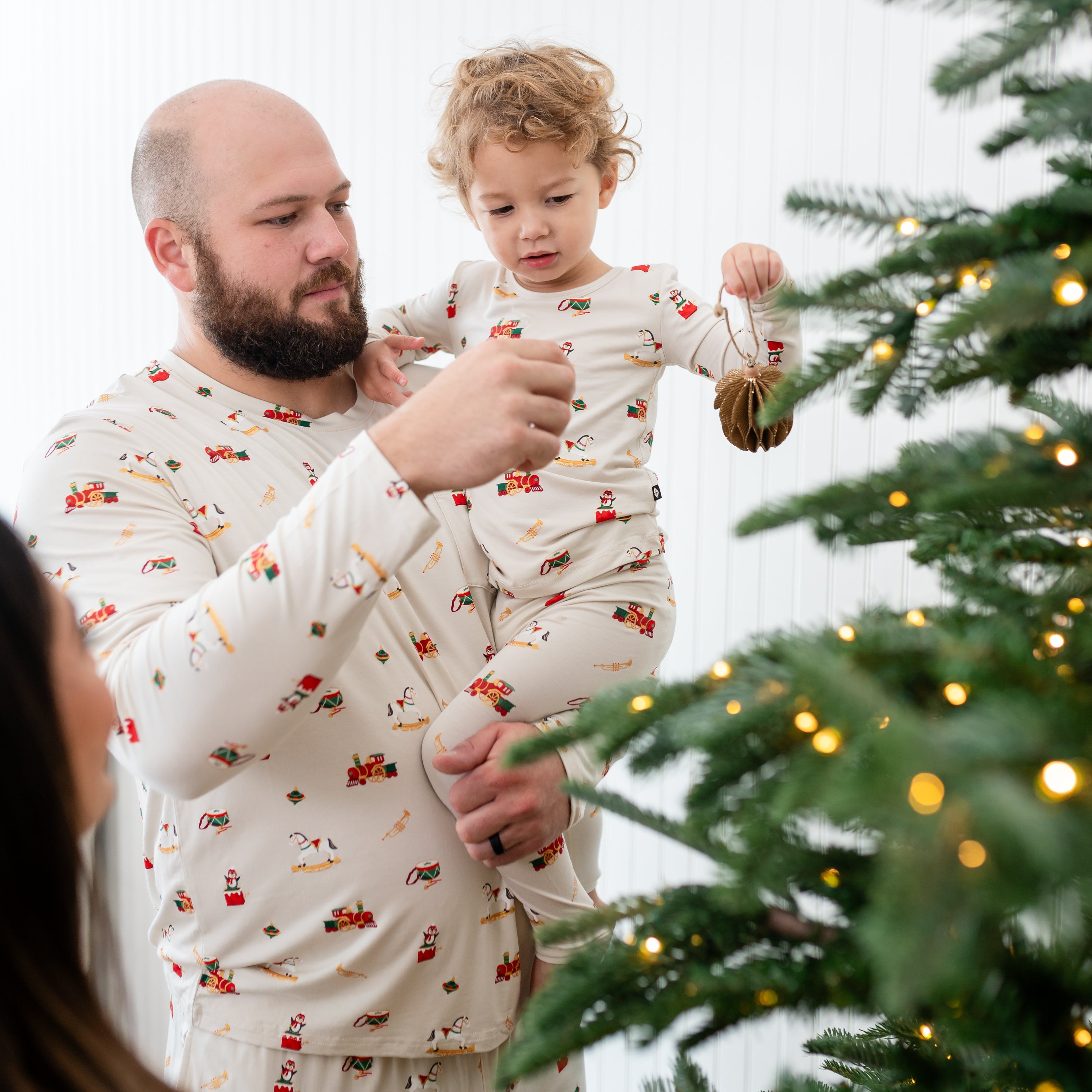 Father holding son who is putting a christmas ornament on the Christmas tree, both matching in Vintage Toys pajamas