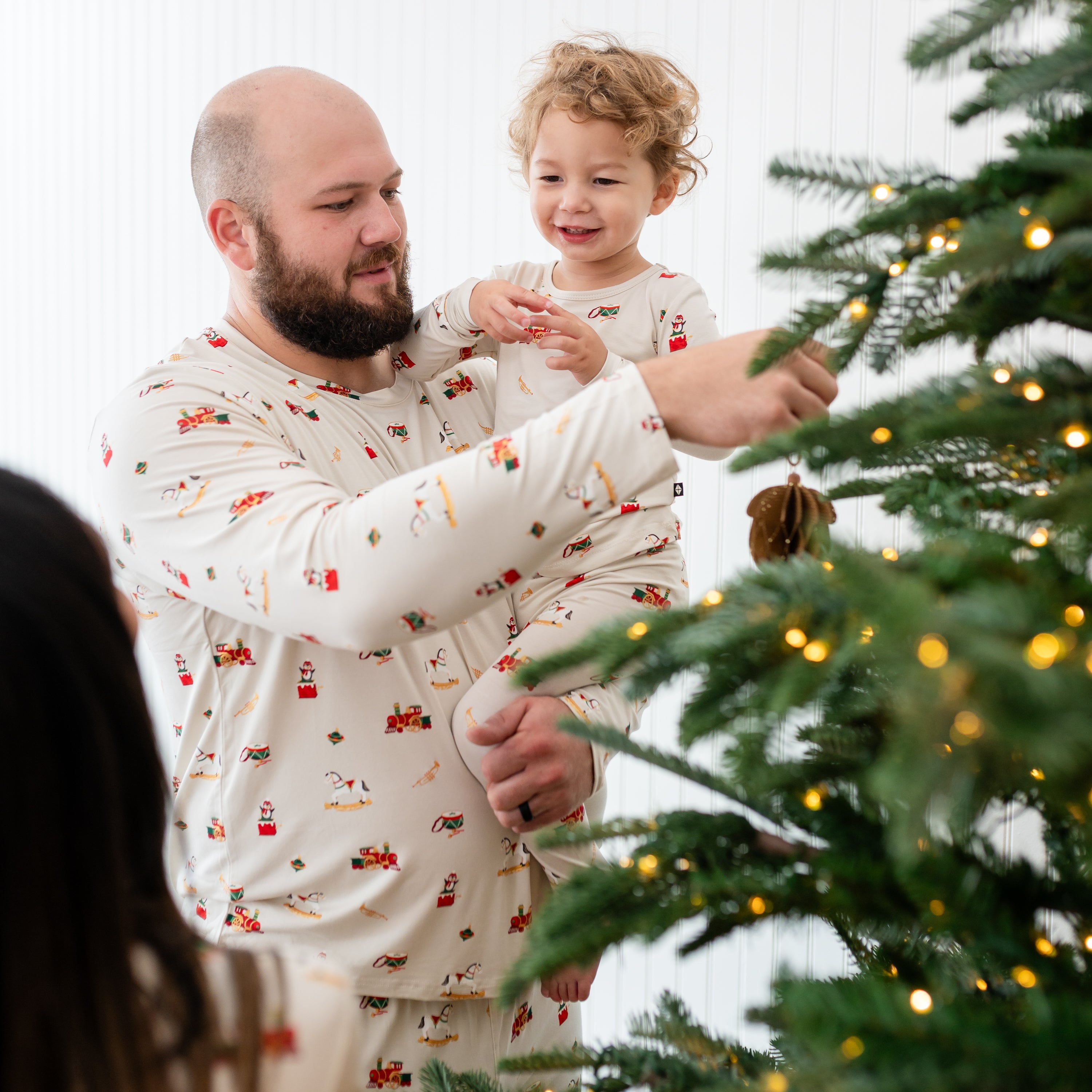 Father wearing the Men's Jogger Set in Vintage Toys holding his son who is matching in long sleeve toddler pajamas putting up a Christmas ornament on a Christmas tree