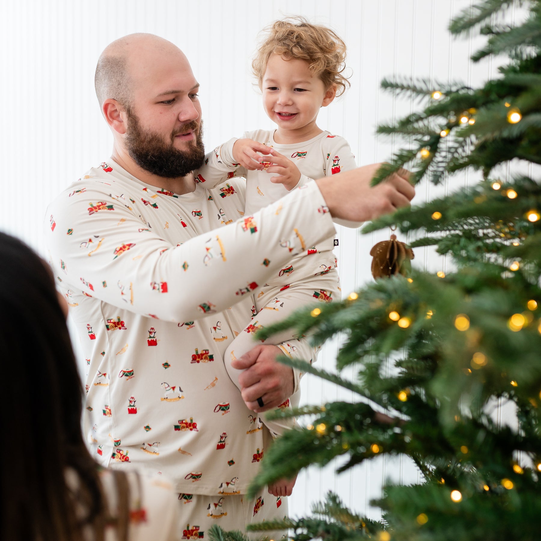 Father wearing the Men's Jogger Set in Vintage Toys holding his son who is matching in long sleeve toddler pajamas putting up a Christmas ornament on a Christmas tree