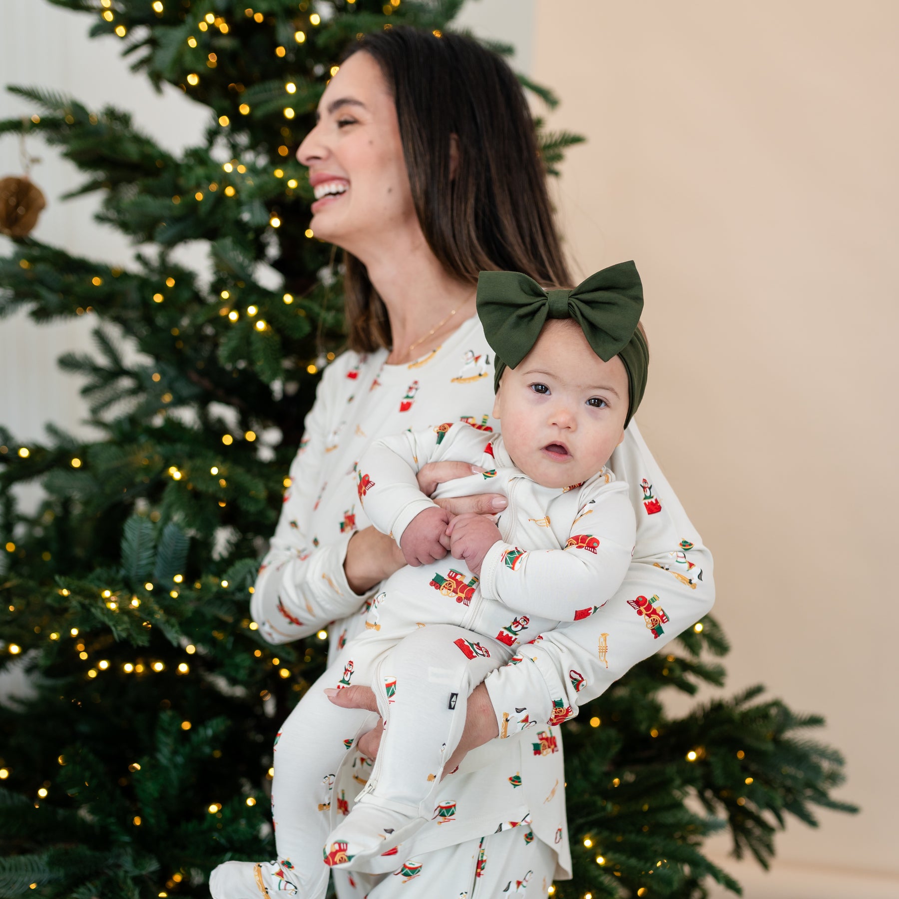 Mother wearing the Women's Jogger Pajama Set in Vintage Toys standing in front of a lit Christmas tree holding her daughter who is wearing a matching zippered footie and Fir Bow Headband