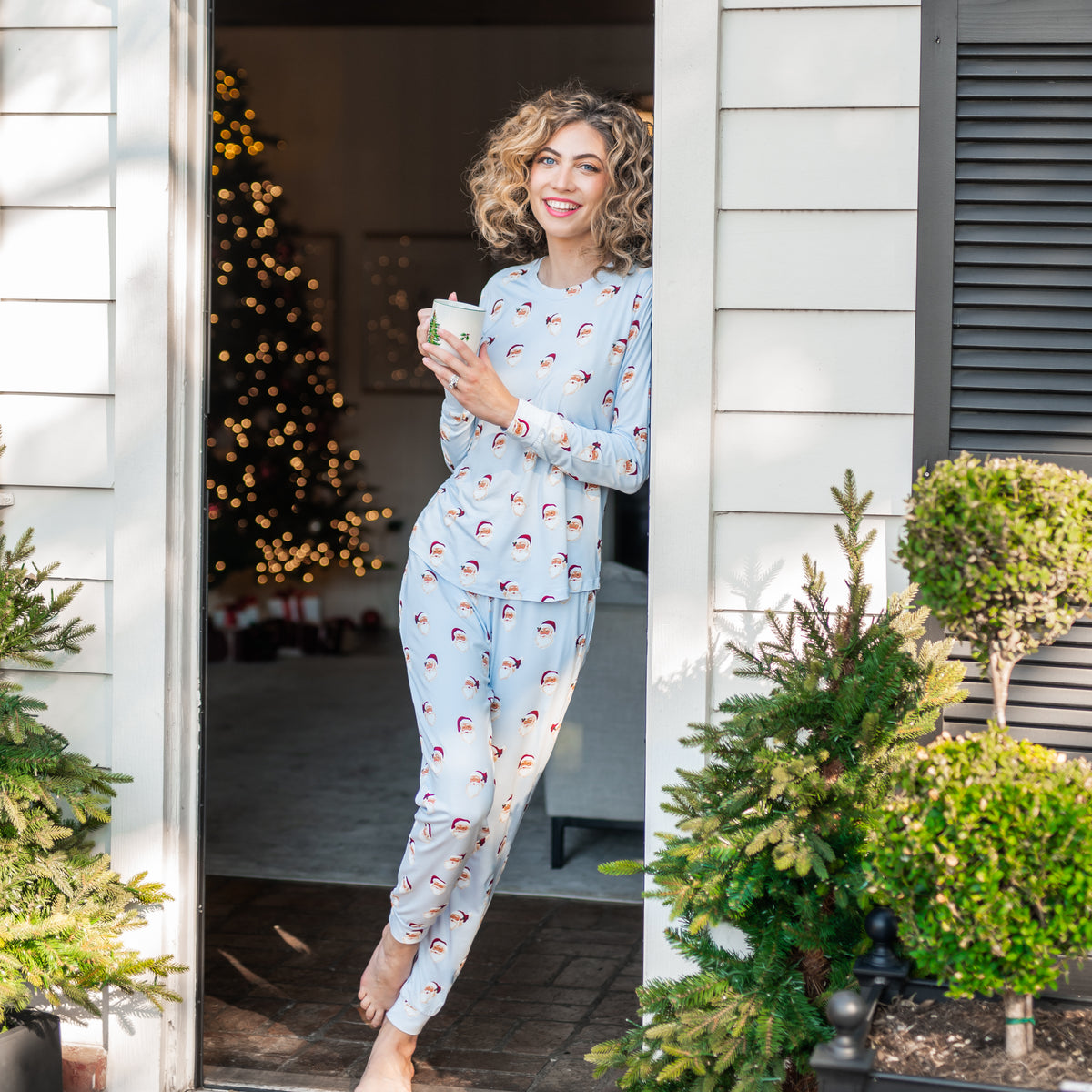 Female model leaning against a door frame on a White House wearing the Women's Jogger Pajama Set in Vintage Santa holding a mug of coffee in her hands