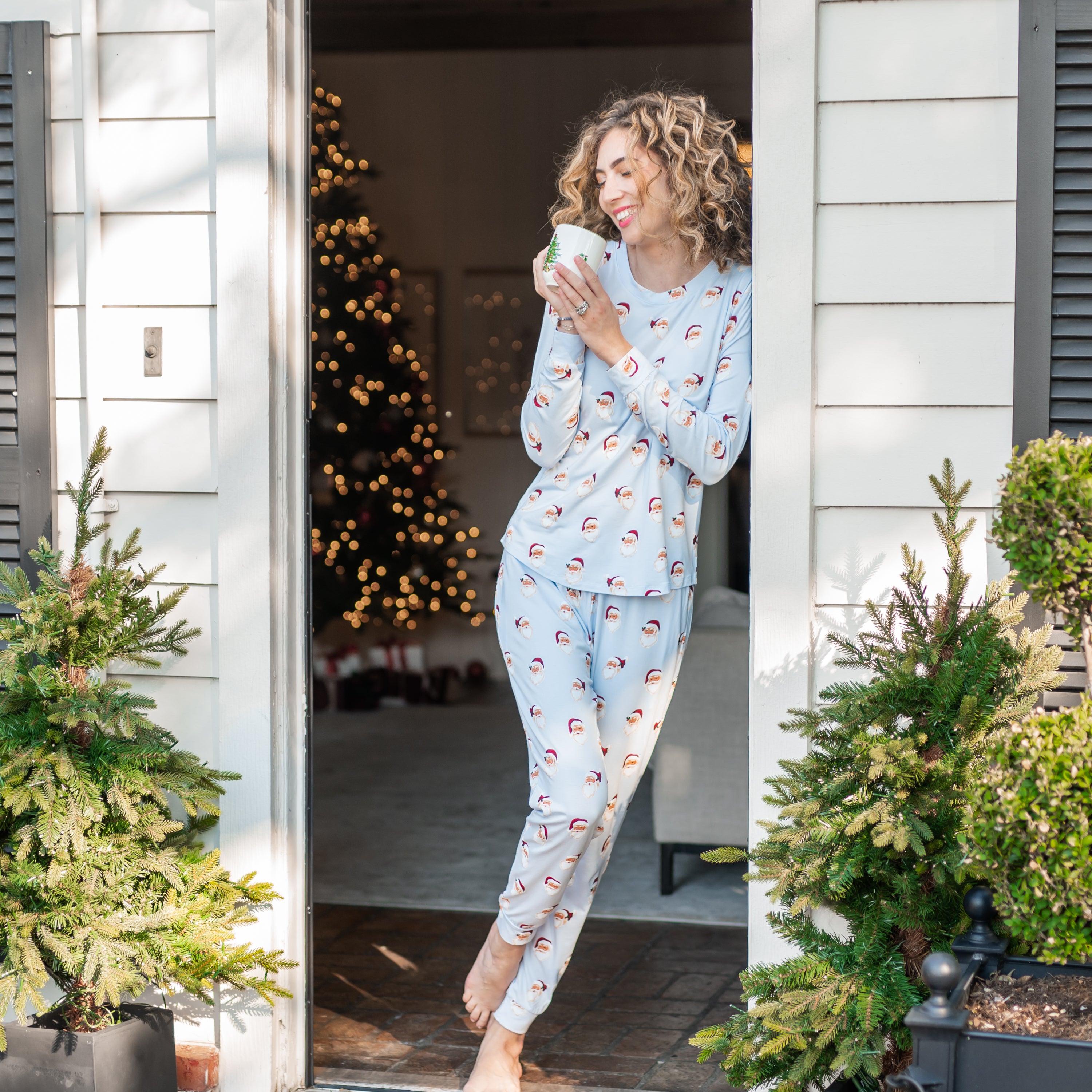 Smiling female model leaning against a door frame to a white house holding a cup of coffee wearing the Women's Jogger Pajama Set in Vintage Santa