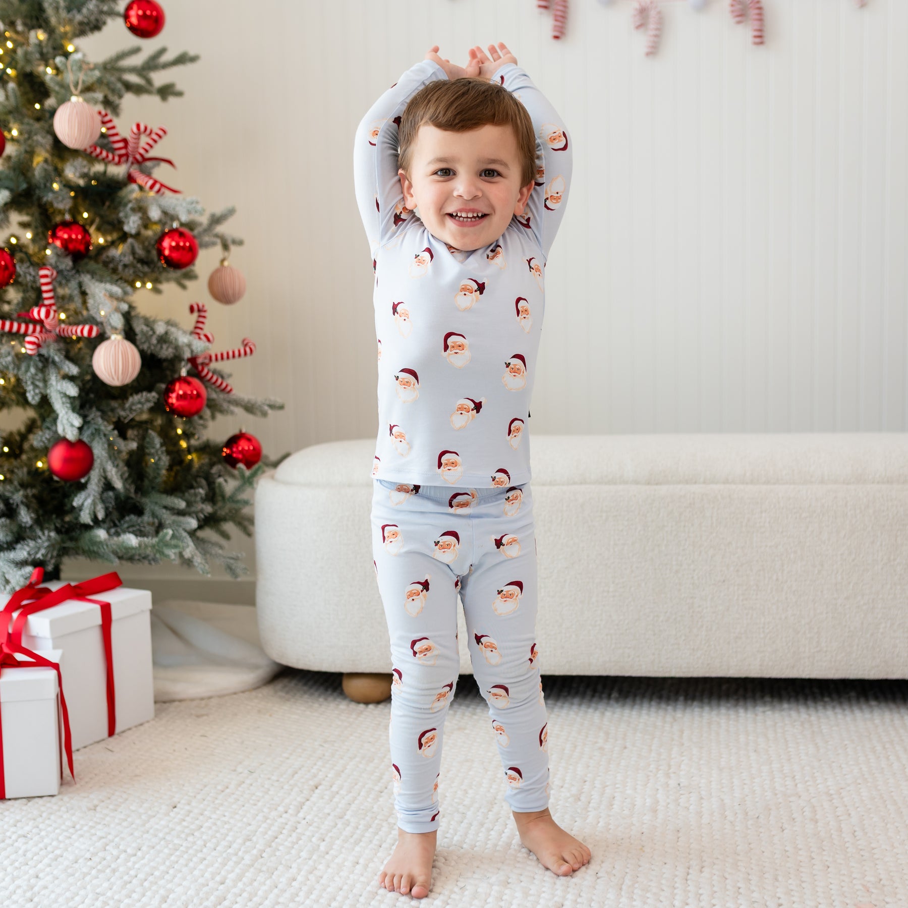 Young toddler standing with his arms in the air wearing the Long Sleeve Pajamas in Vintage Santa in front of a cream ottoman and decorated Christmas tree