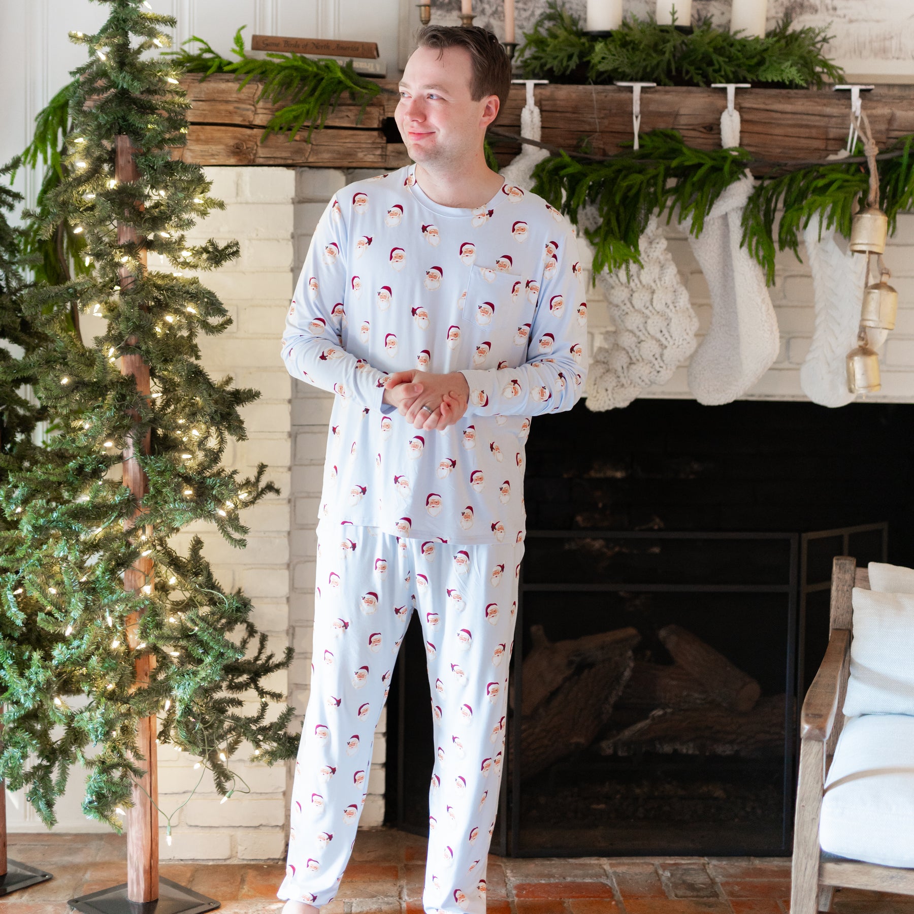 Male model standing with his hands clasped in front of him with a white brick fireplace behind him wearing the Men's Jogger Set in Vintage Santa