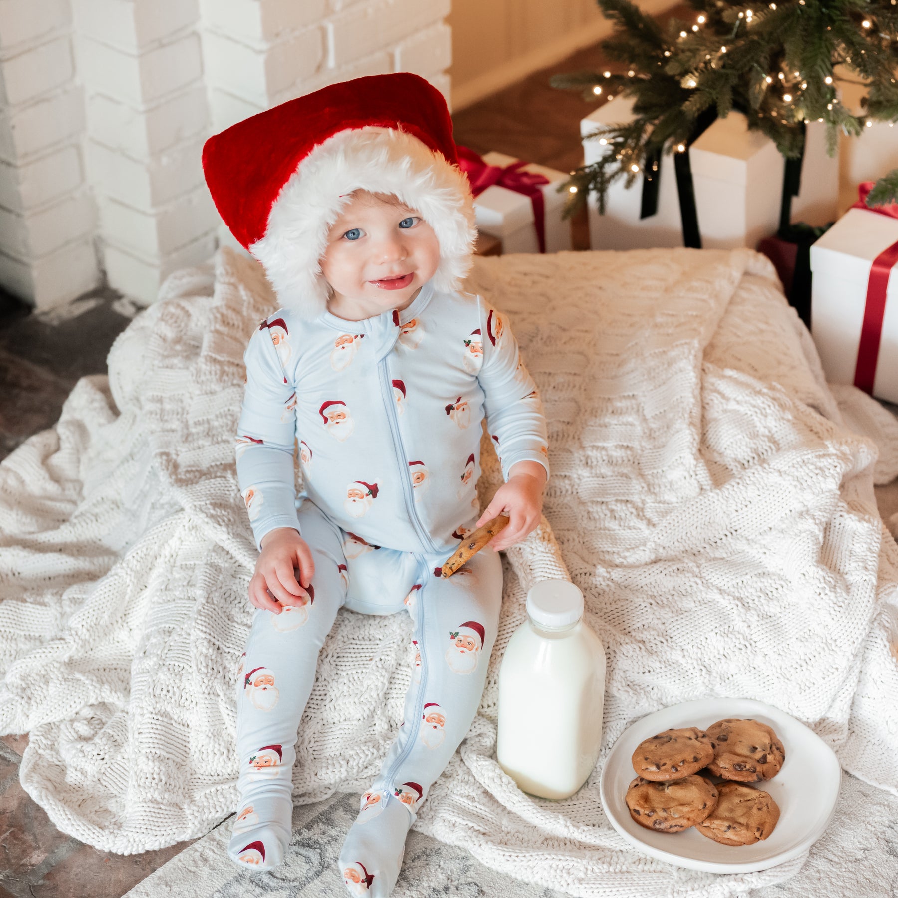 Young toddler sitting on a white blanket beside a jug of milk and a plate of cookies holding a cookie in his hand wearing the Zippered Footie in Vintage Santa with a red and white Santa Hat