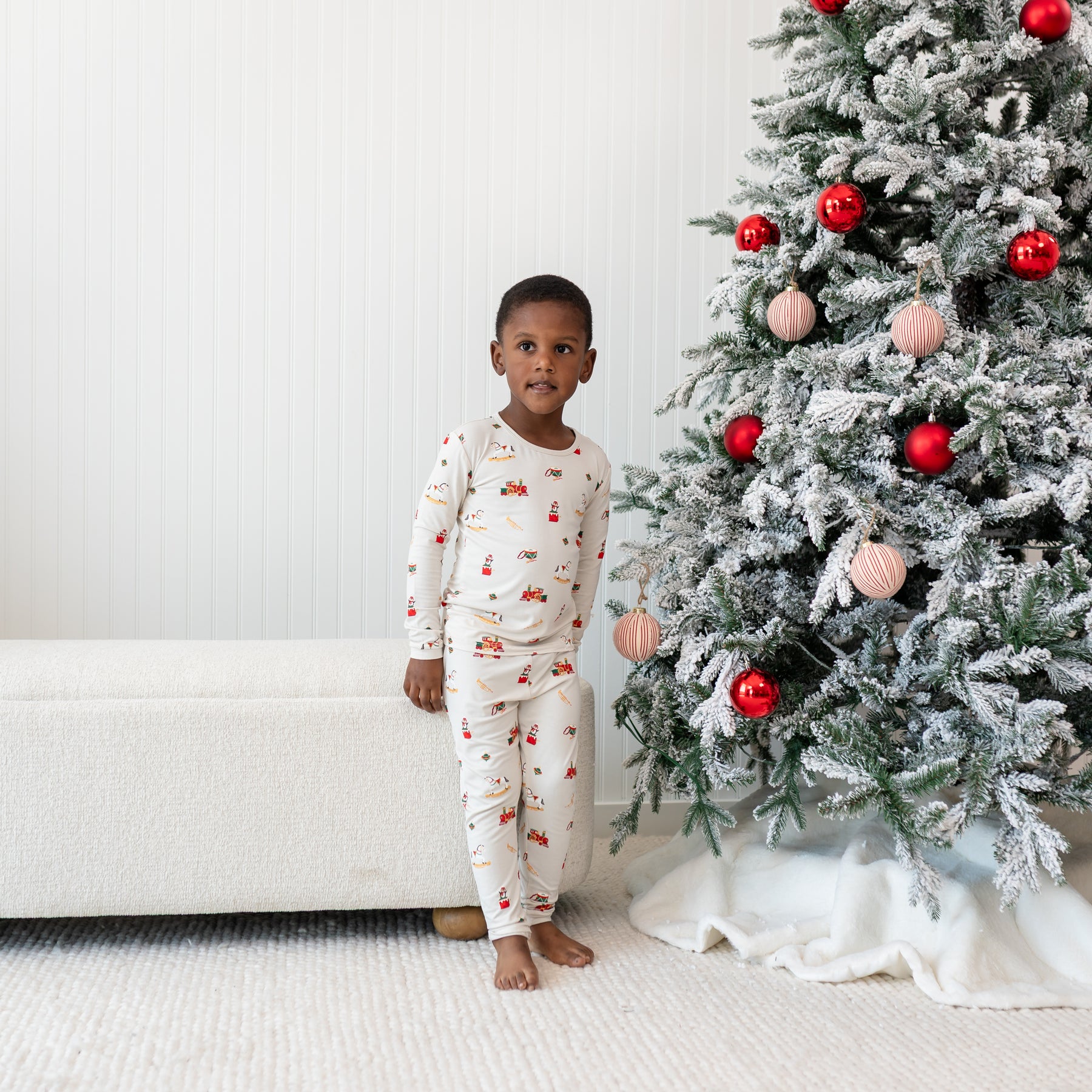 Young boy standing in front of a cloth bench beside a decorated Christmas Tree wearing the Long Sleeve Pajamas in Vintage Toys