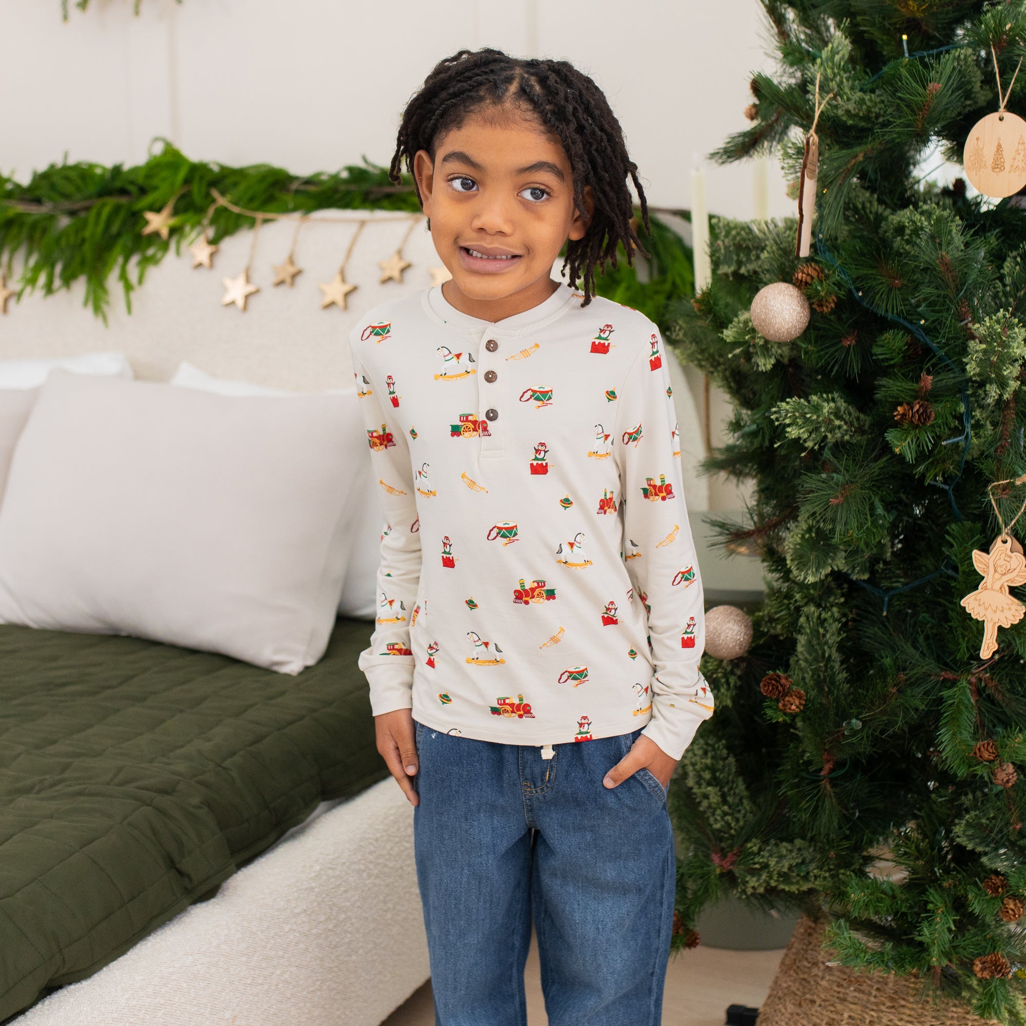 Young girl standing between a decorated Christmas tree and bed wearing the Long Sleeve Toddler Henley Tee in Vintage Toys with one hand in her jeans pocket