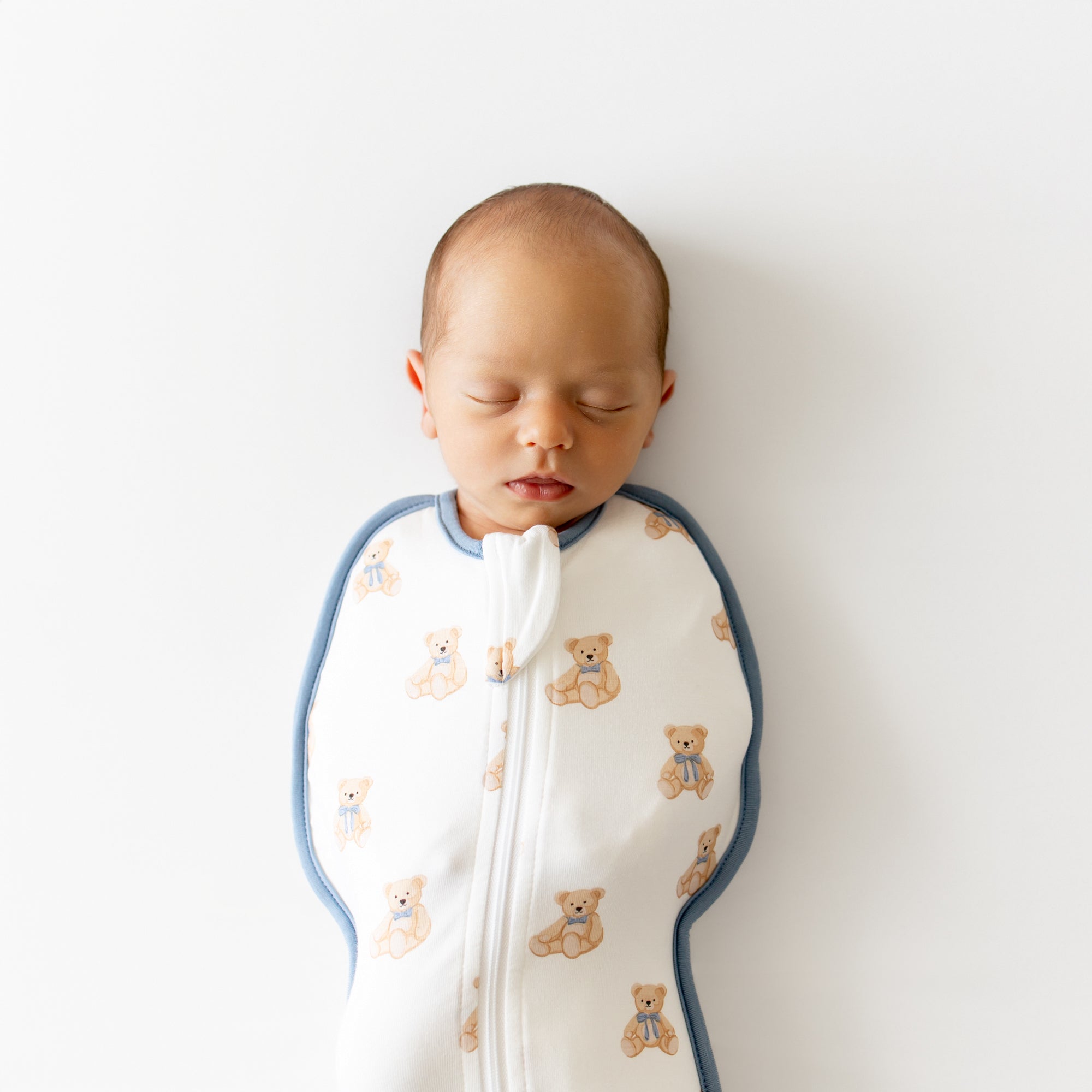 Newborn baby sleeping peacefully in a white swaddle with teddy bear pattern on a light background