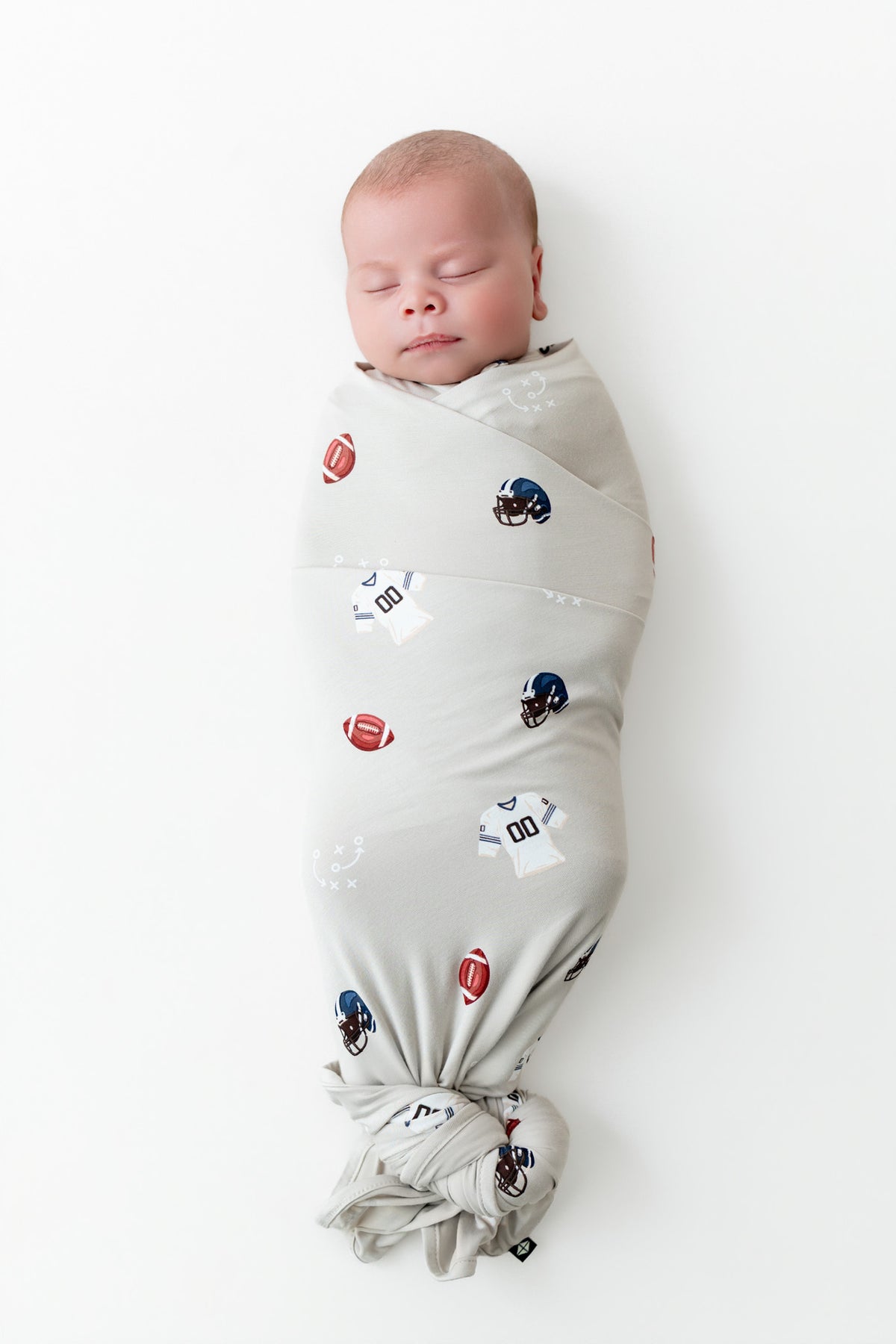 A sleeping newborn baby swaddled in a light gray fabric swaddle blanket with a pattern of footballs and helmets, photographed against a clean white background.
