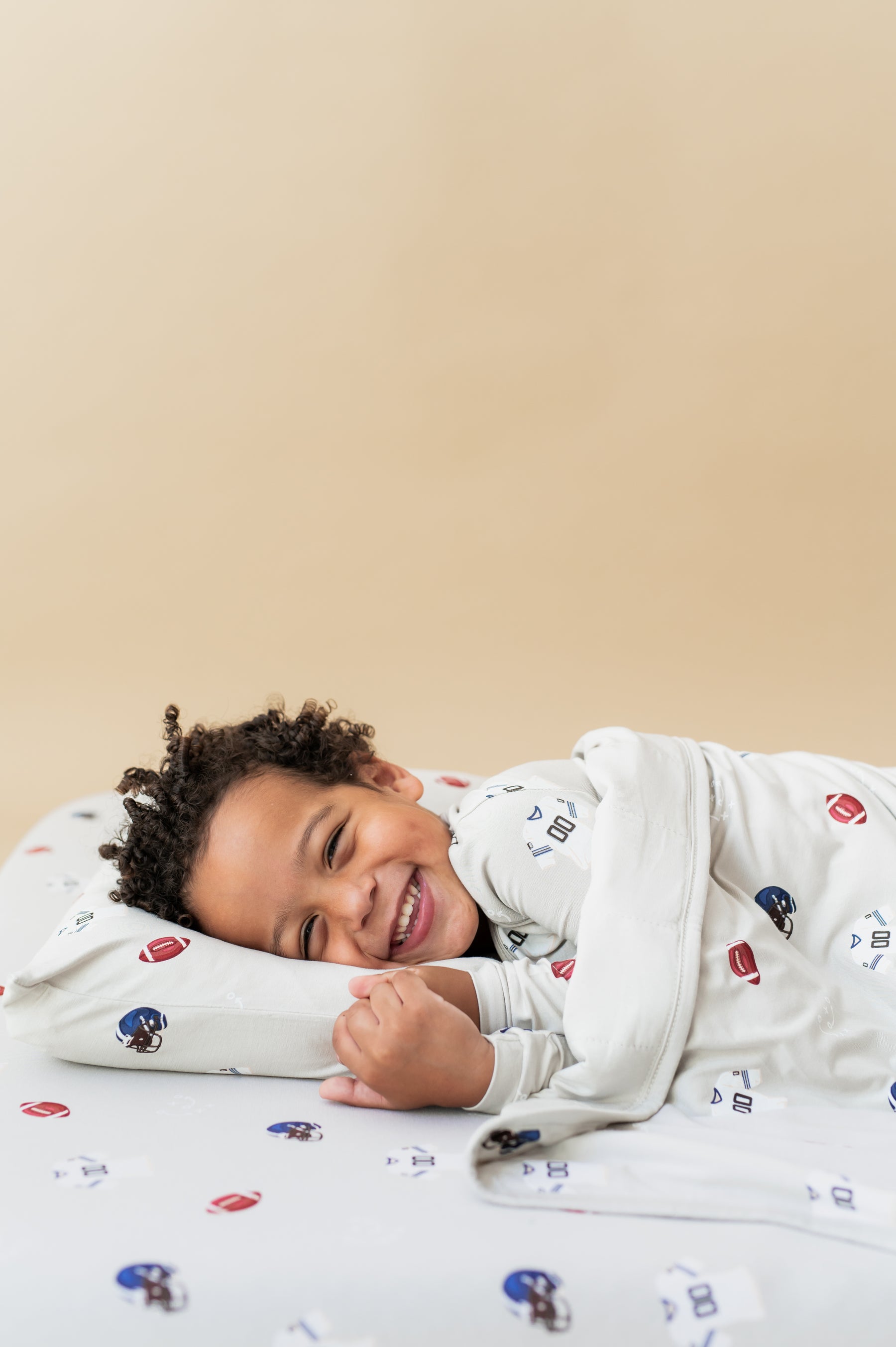 Boy lying on Toddler Pillowcase and sheet in Vintage Football print while wearing a similar print long sleeve pajama set covered with a similar print toddler blanket on a beige background