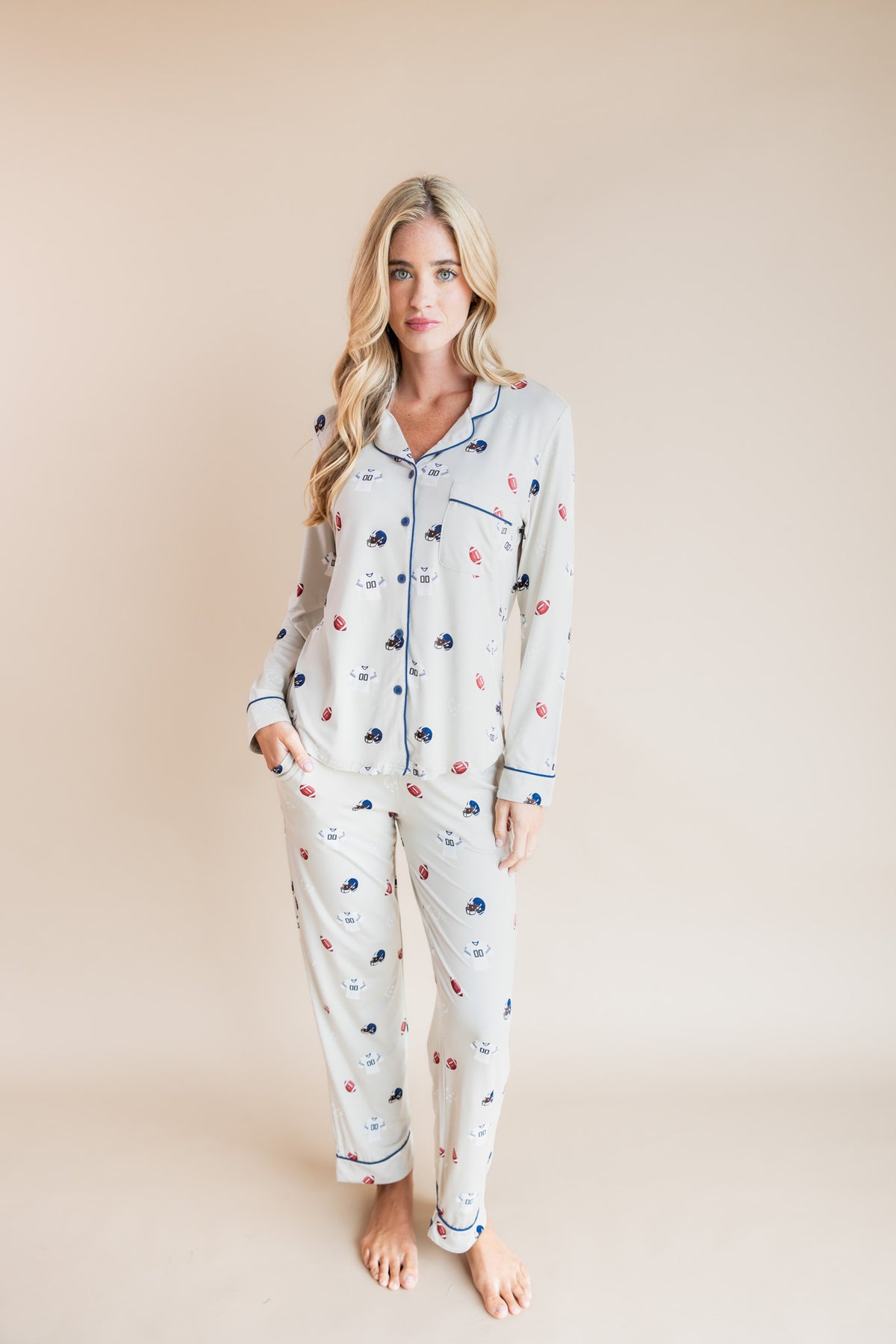 A woman modeling a two-piece pajama set with a light gray base and a small multicolor pattern of football motifs, paired with solid blue piping, set against a solid beige background.