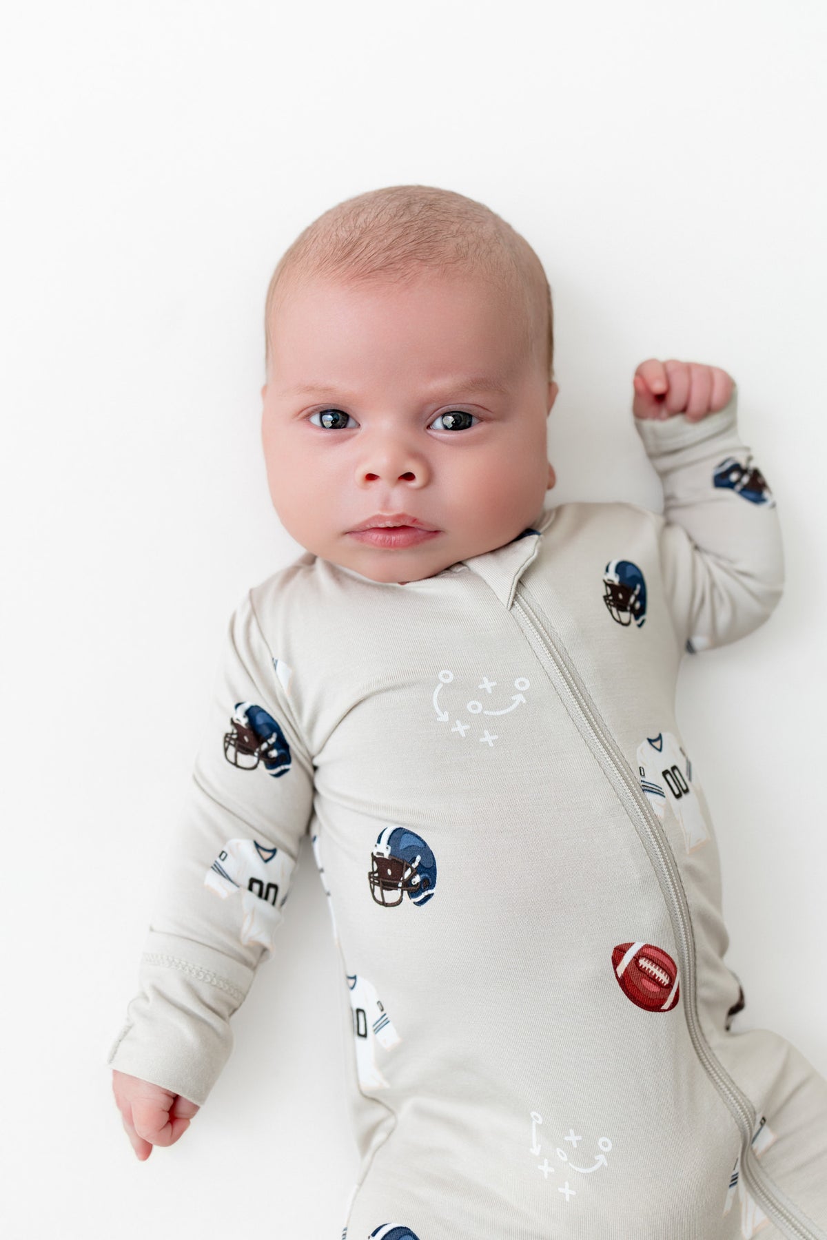 A baby lying on their back wearing a light beige long-sleeve zip-up sleeper with a pattern of football helmets and a football.