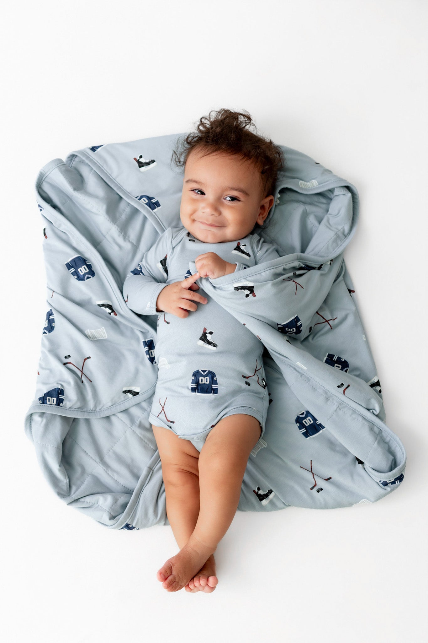 Baby smiling over Baby Blanket in Vintage Hockey 1.0 print while wearing a similar print bodysuit on a white background