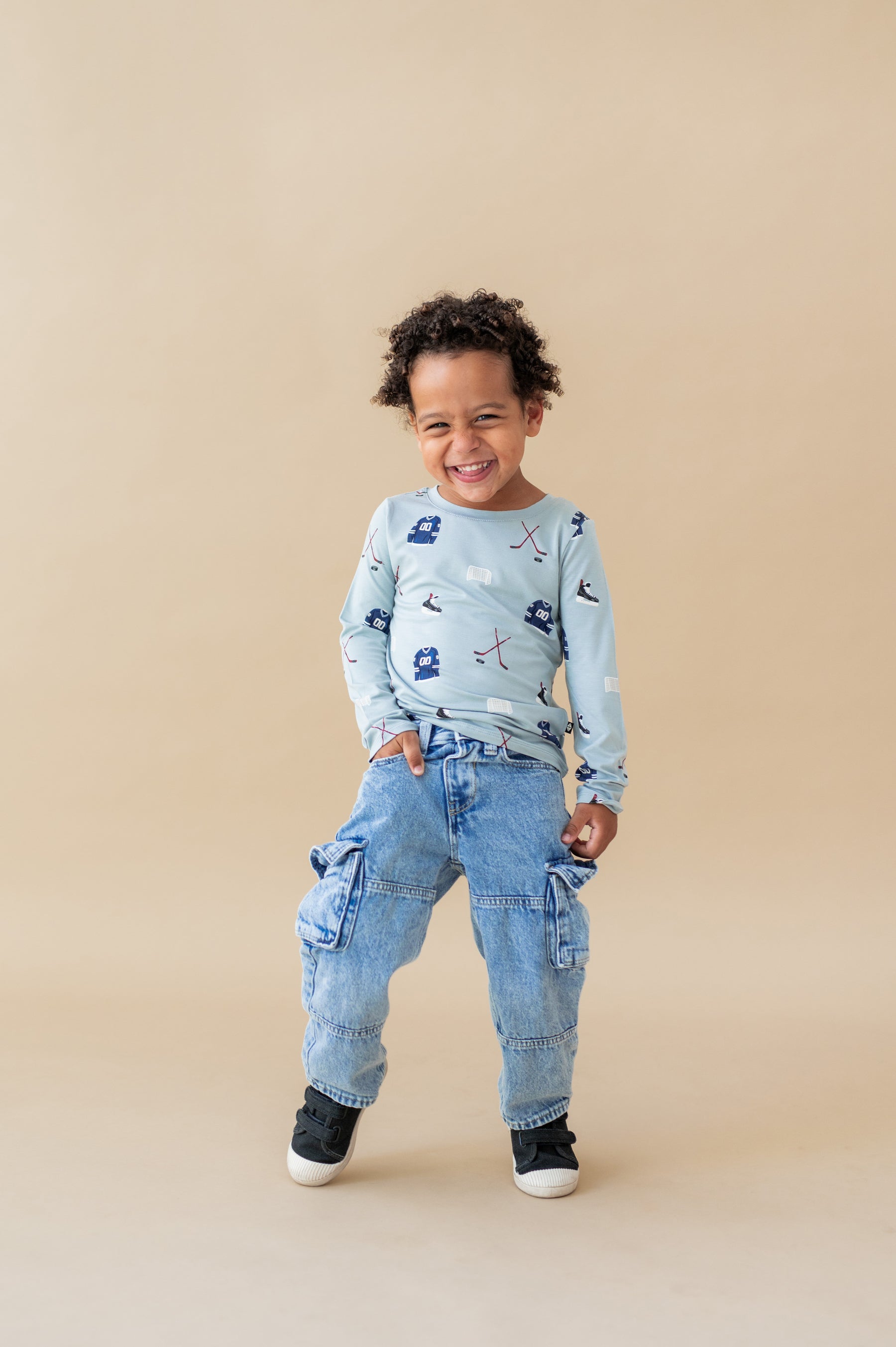 Boy wearing Long Sleeve Toddler Crew Neck Tee in Vintage Hockey print over blue jeans and black shoes on a beige background
