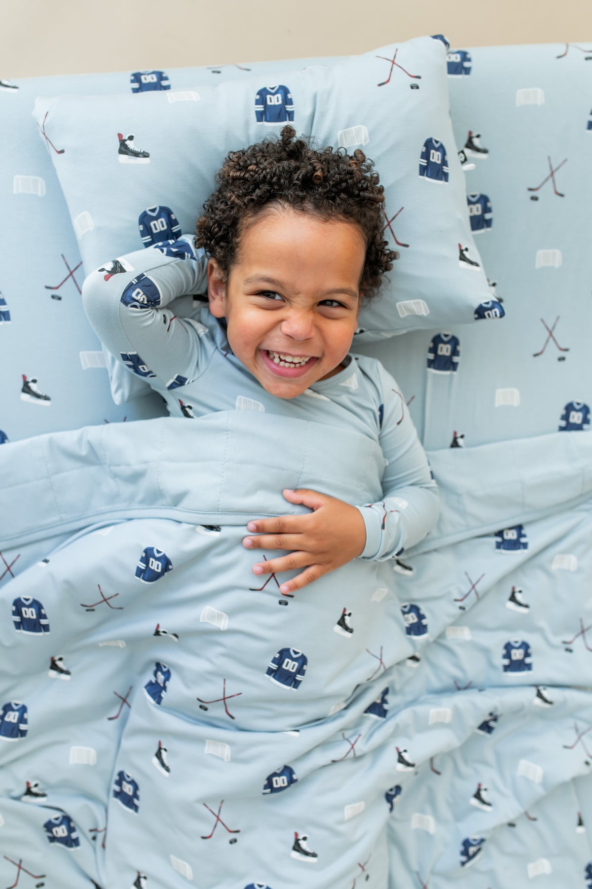 Boy lying on Toddler Pillowcase in Vintage Hockey print while wearing a similar print long sleeve pajama set smiling on a similar print sheet and covered with similar print blanket