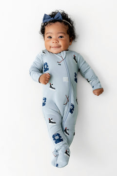 Baby wearing Zippered Footie in Vintage Hockey print and blue bow headband on a white background