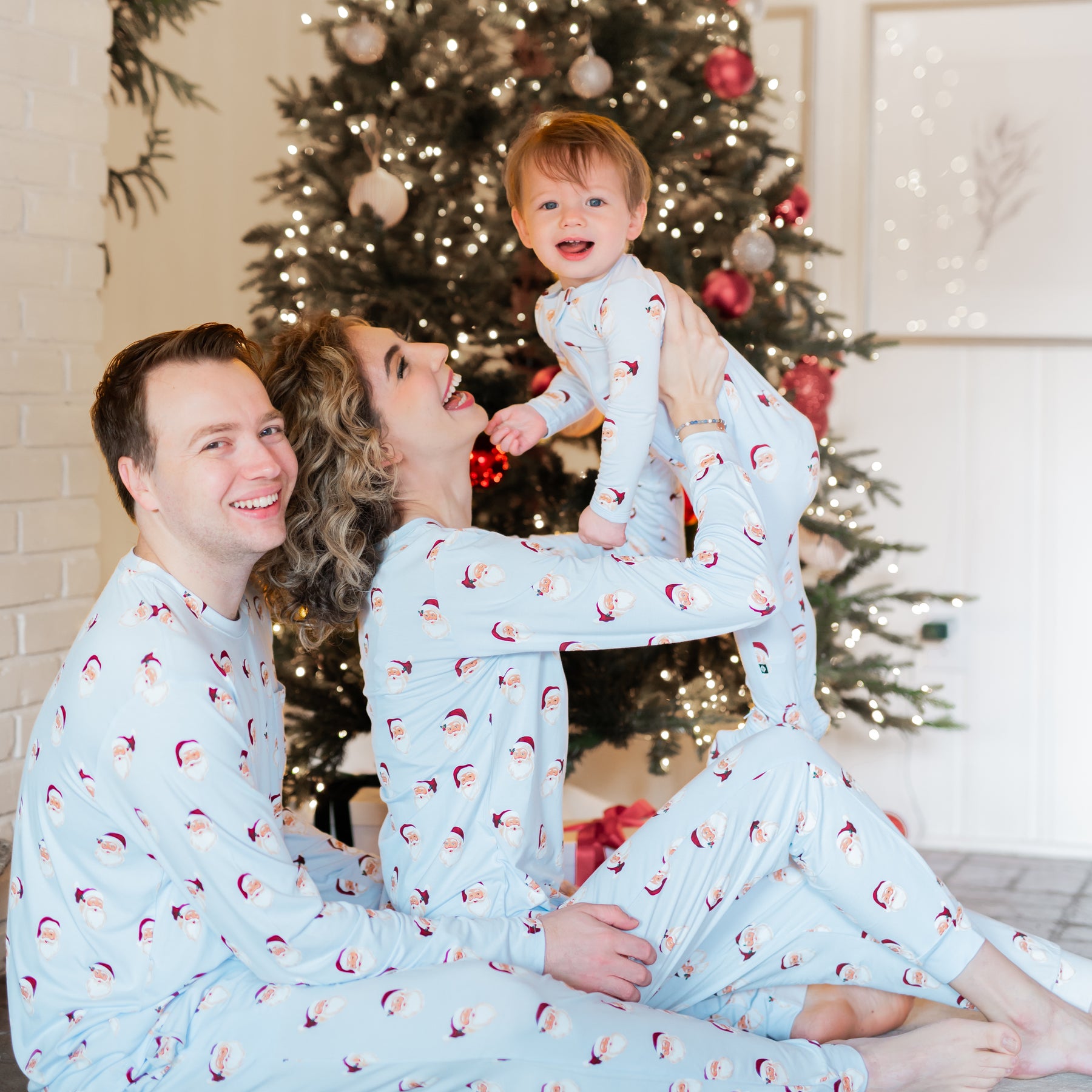 Family of three sitting on the floor in front of a Christmas tree matching in Vintage Santa. Mother is holding son up in the air with her husband sitting beside her