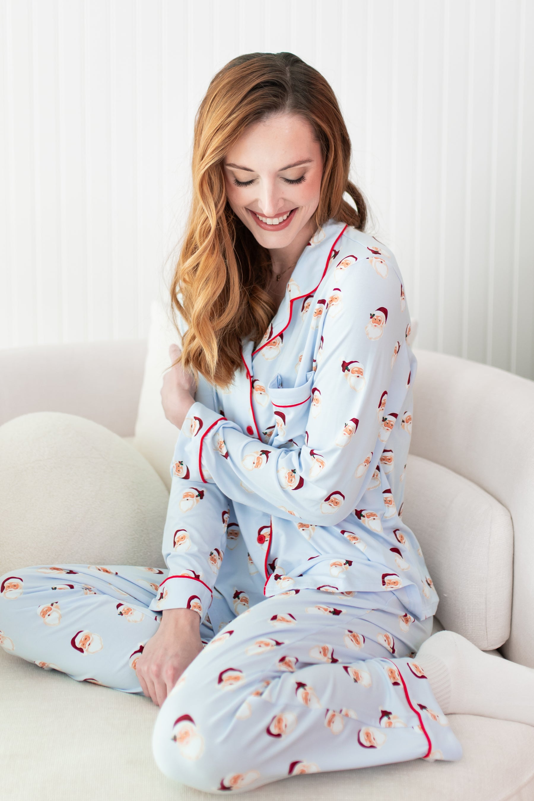 Female model wearing the Long-Sleeved Women's Pajama Set in Vintage Santa sitting on a cream color sofa