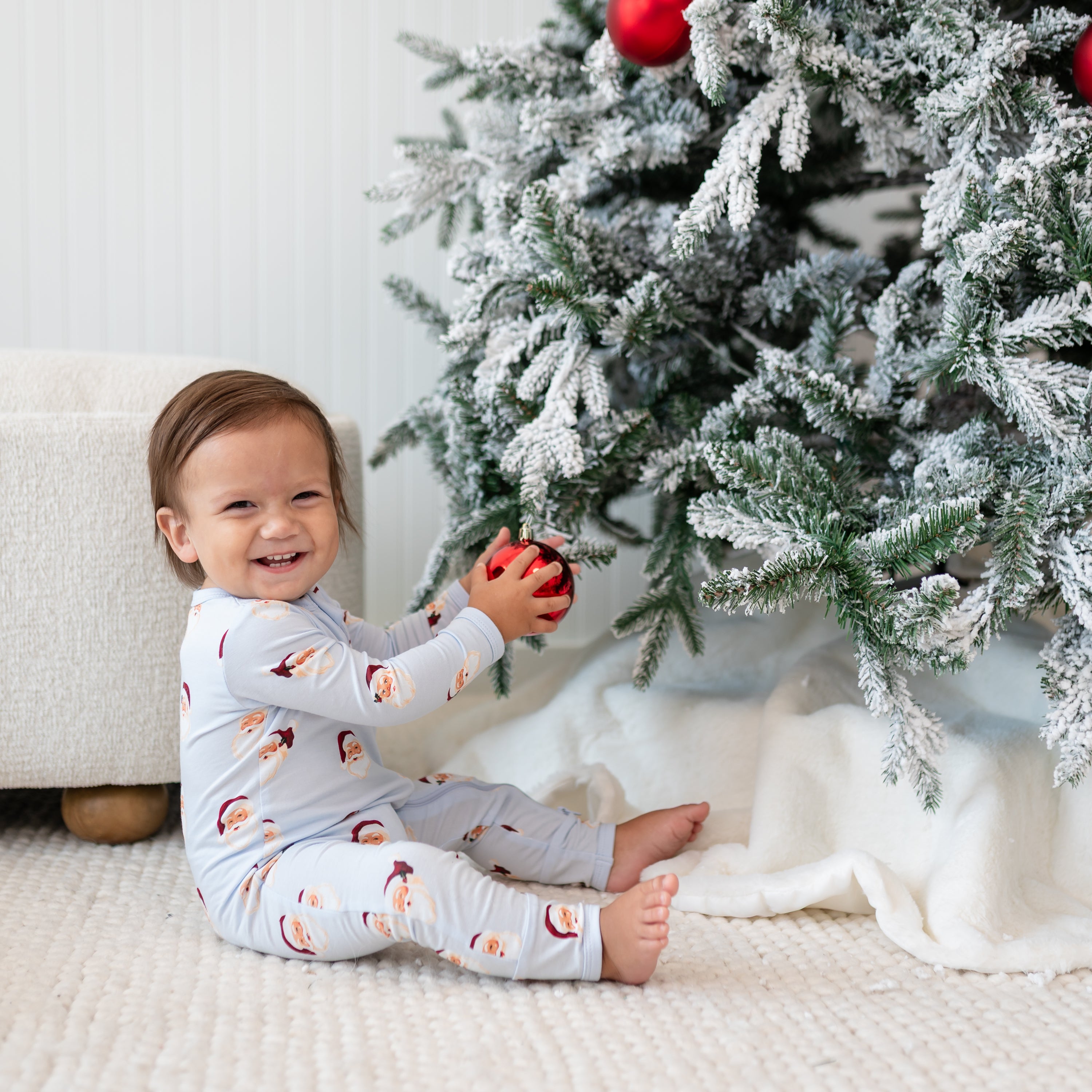 Smiling toddler sitting beside a christmas tree wearing the Zippered Romper in Vintage Santa holding an ornament