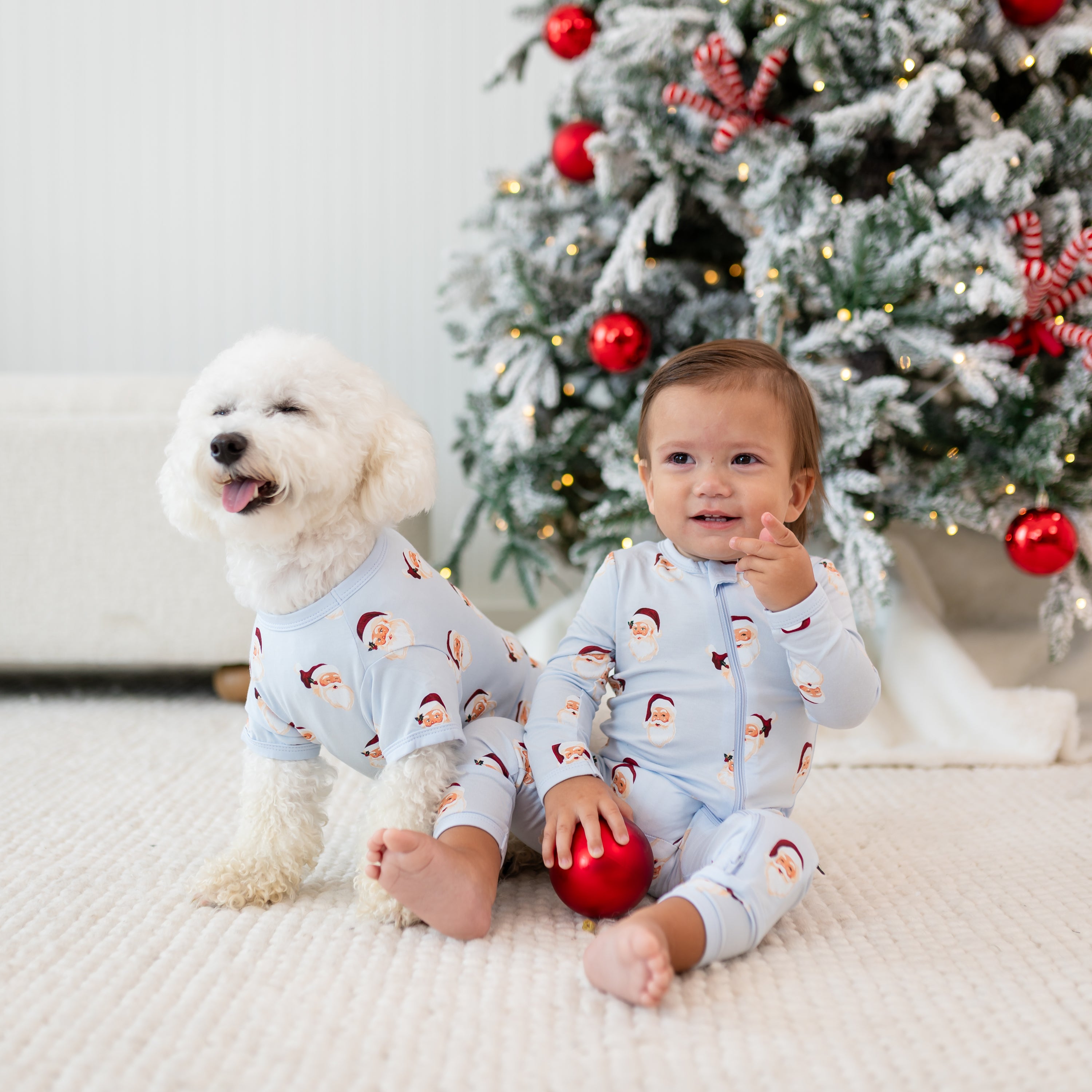 Small white dog wearing the Dog Tee in Vintage Santa sitting on the floor beside a young toddler who is wearing a matching zippered romper