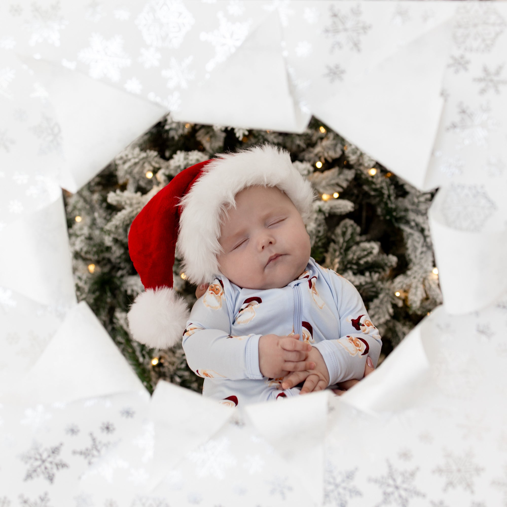 Sleeping infant wearing the Zippered Romper in Vintage Santa and a santa hat in front of a christmas tree through the view of a hole in white snowflake wrapping paper