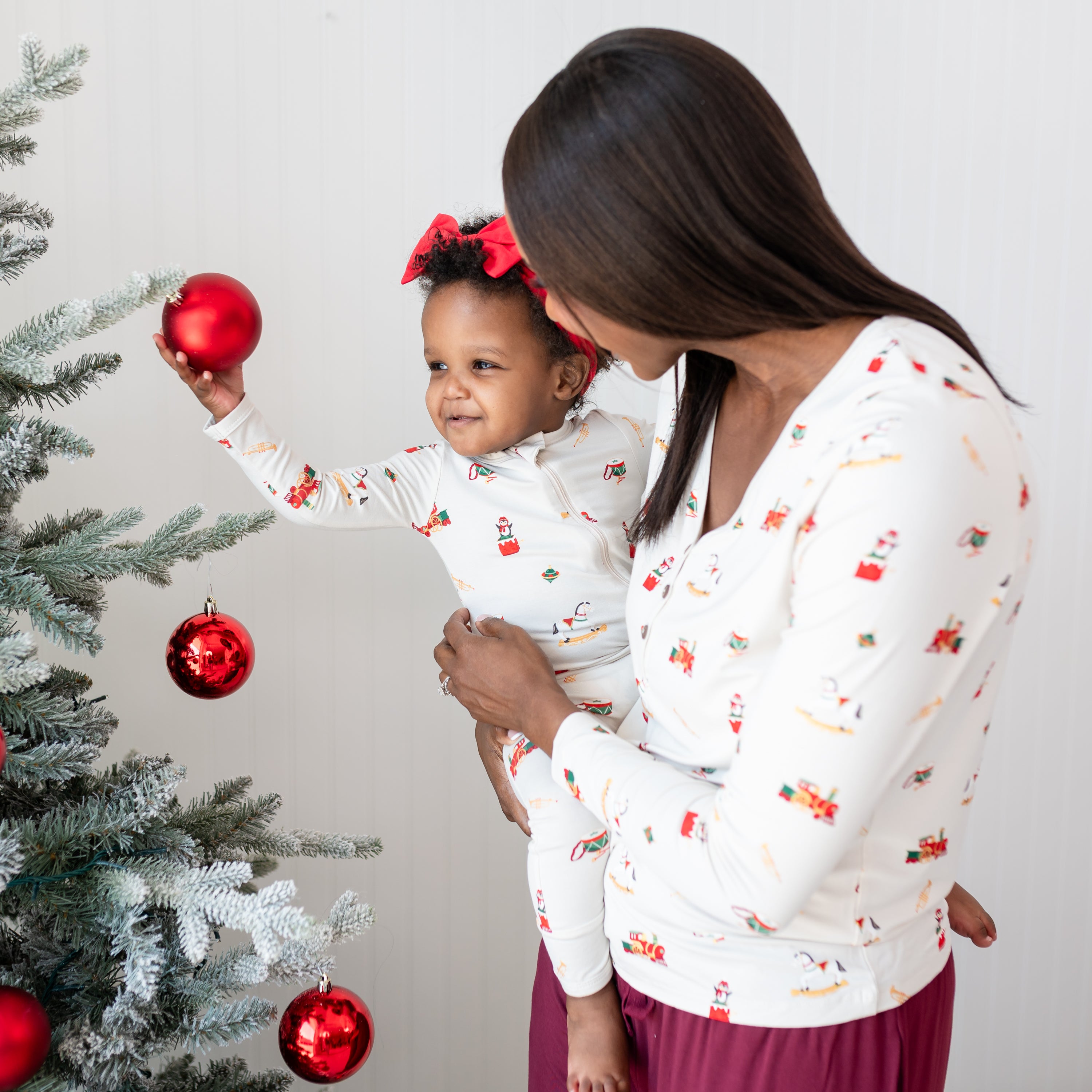 Mother wearing the Women's Henley Tee in Vintage Toys, holding her daughter who is wearing a matching Zippered Romper in Vintage Toys who is touching a red Christmas ball ornament on a Christmas tree