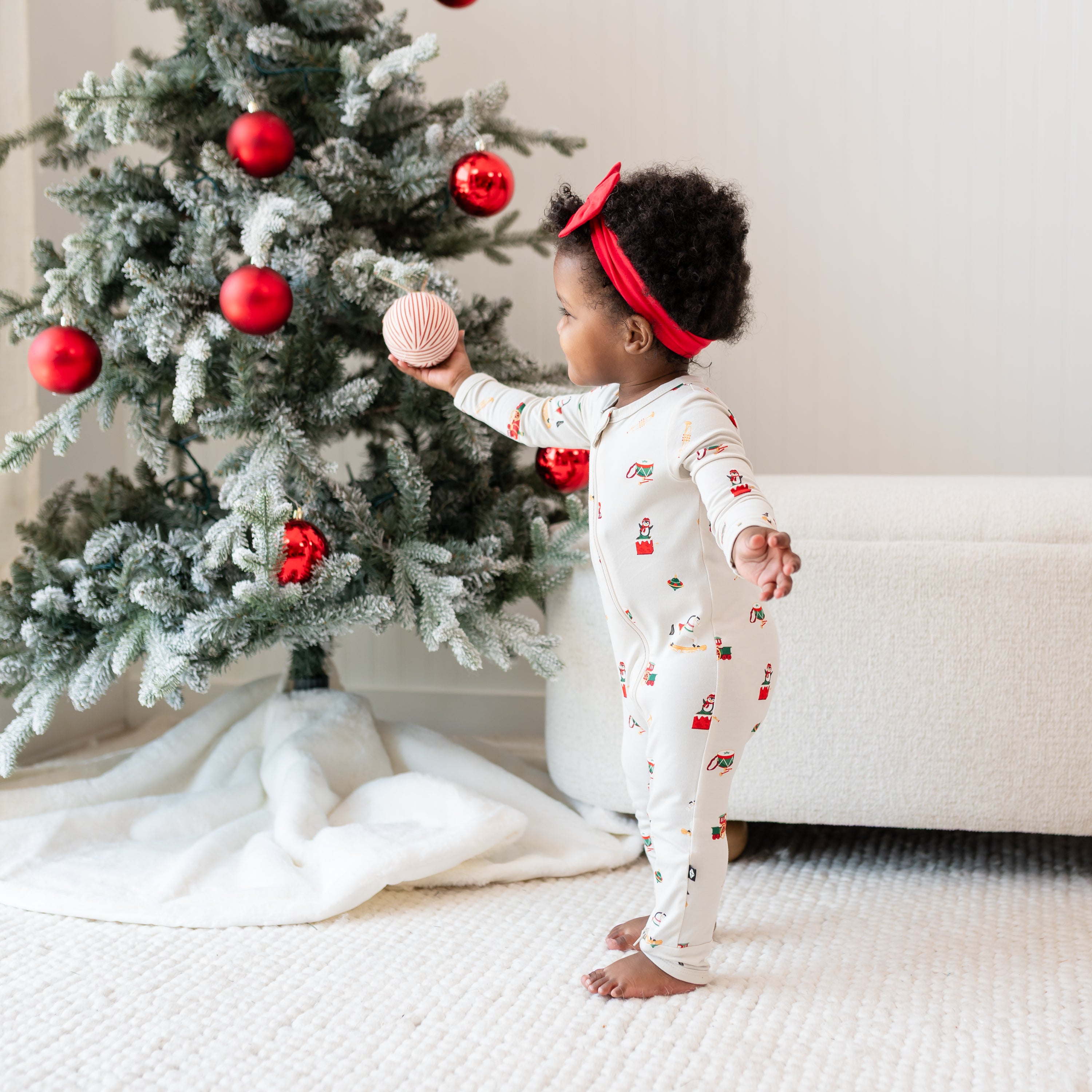 Young toddler wearing the Zippered Romper in Vintage Toys standing in front of a decorated Christmas tree holding a red and white striped ball decoration