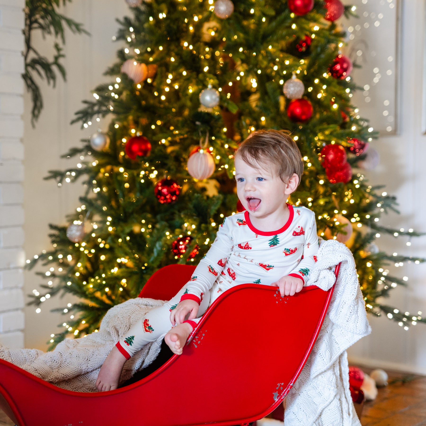 Young boy sitting in a red sleigh in front of a decorated Christmas tree wearing the Long Sleeve Pajamas in Santa Sleigh