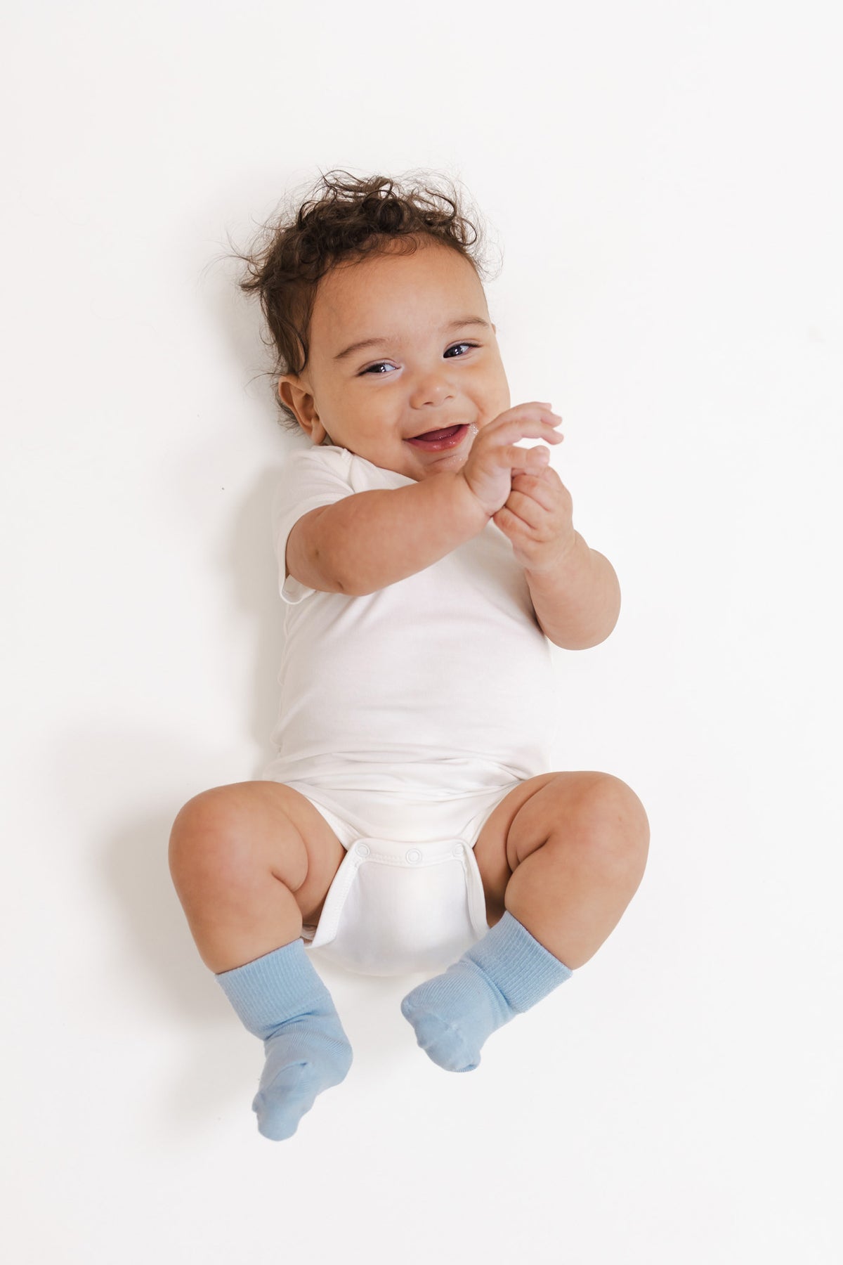 A smiling baby lying on a white background, wearing a white short-sleeve bodysuit and light blue socks.