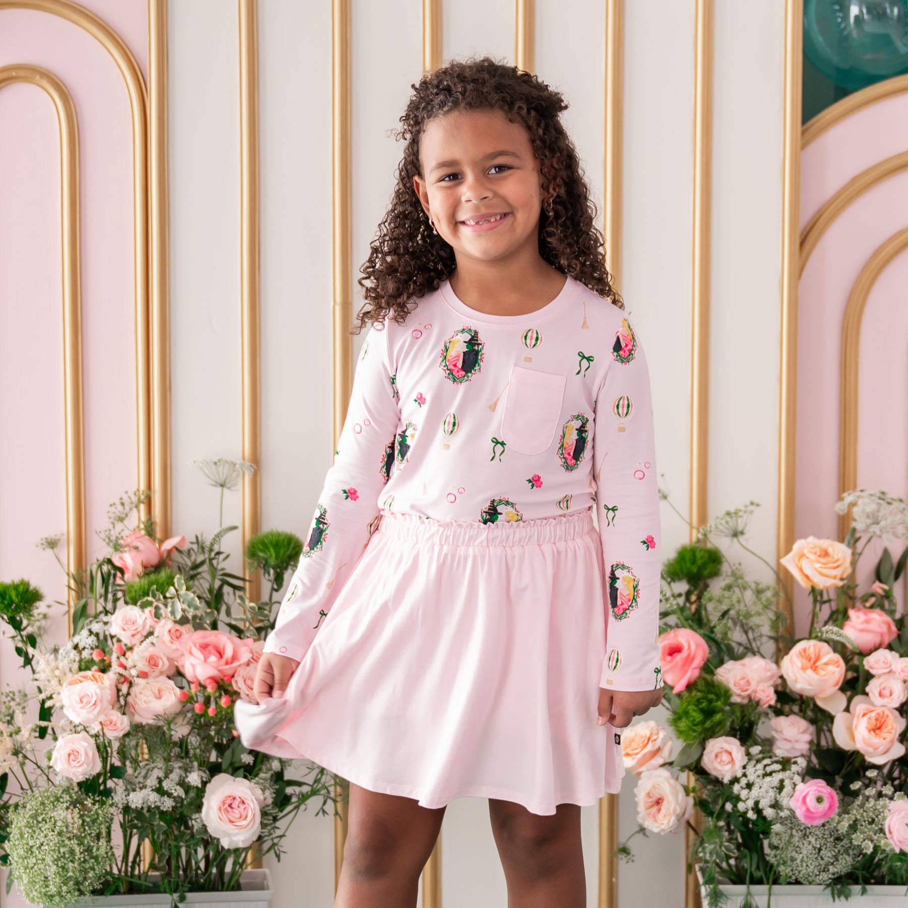 Smiling girl wearing the soft and breathable Long Sleeve Toddler Crew Neck Tee in Wicked from Kyte Baby paired with a Sakura Skort standing in front of a floral backdrop