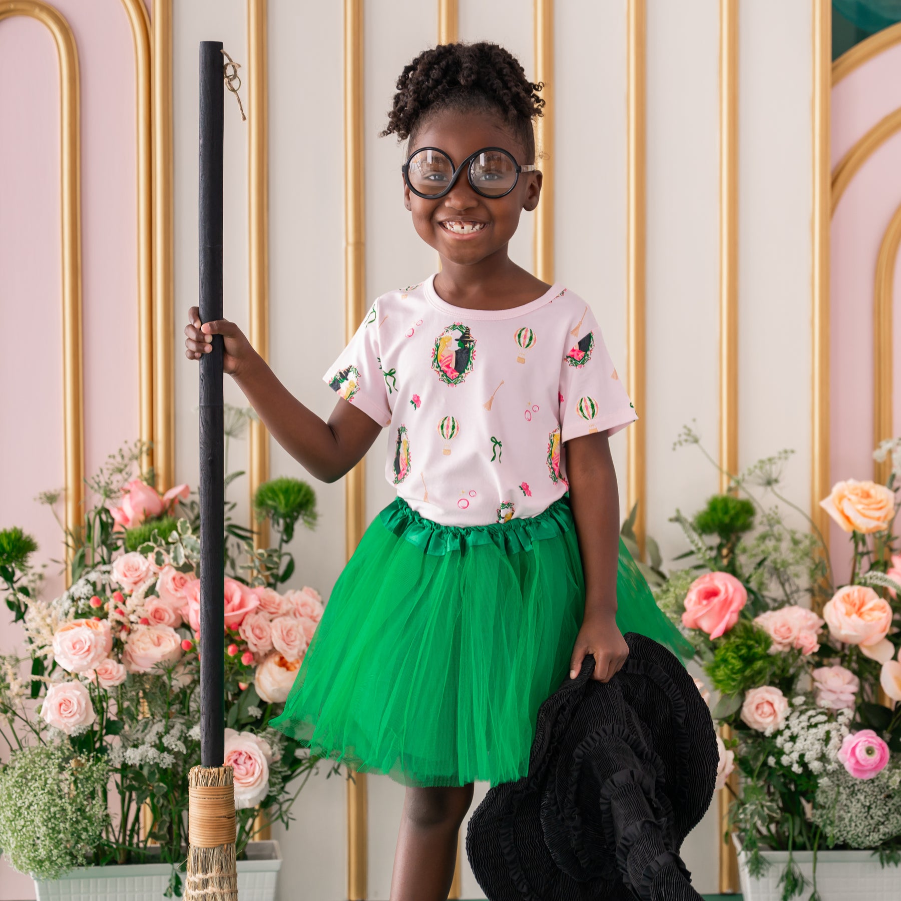 Smiling girl standing holding a broom and black witches hat wearing circle prop glasses and Toddler Basic Tee in Wicked with a green tulle skirt