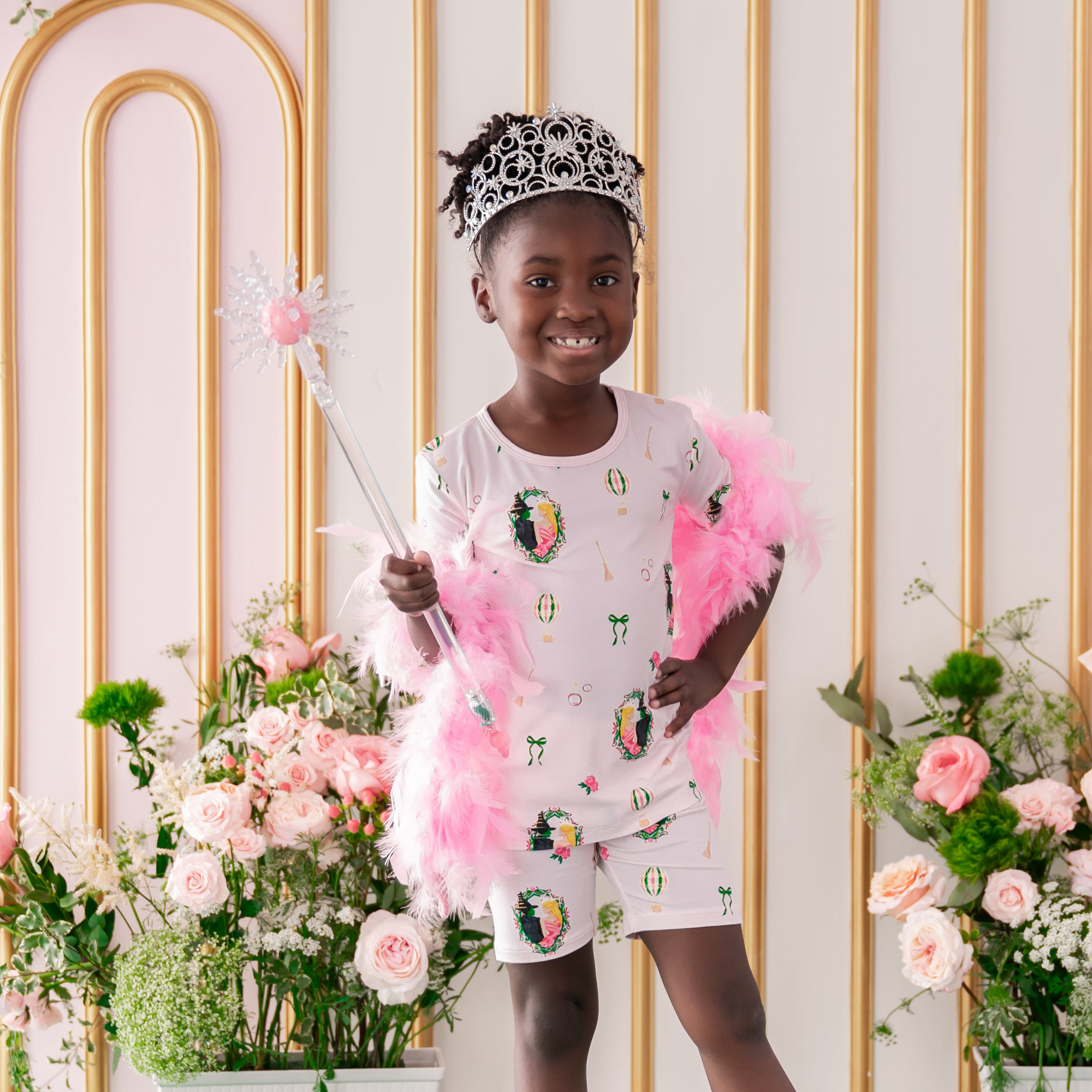 Young girl wearing the Short Sleeve Pajamas in Wicked, a play tiara and a wand standing in front of a flower backdrop 