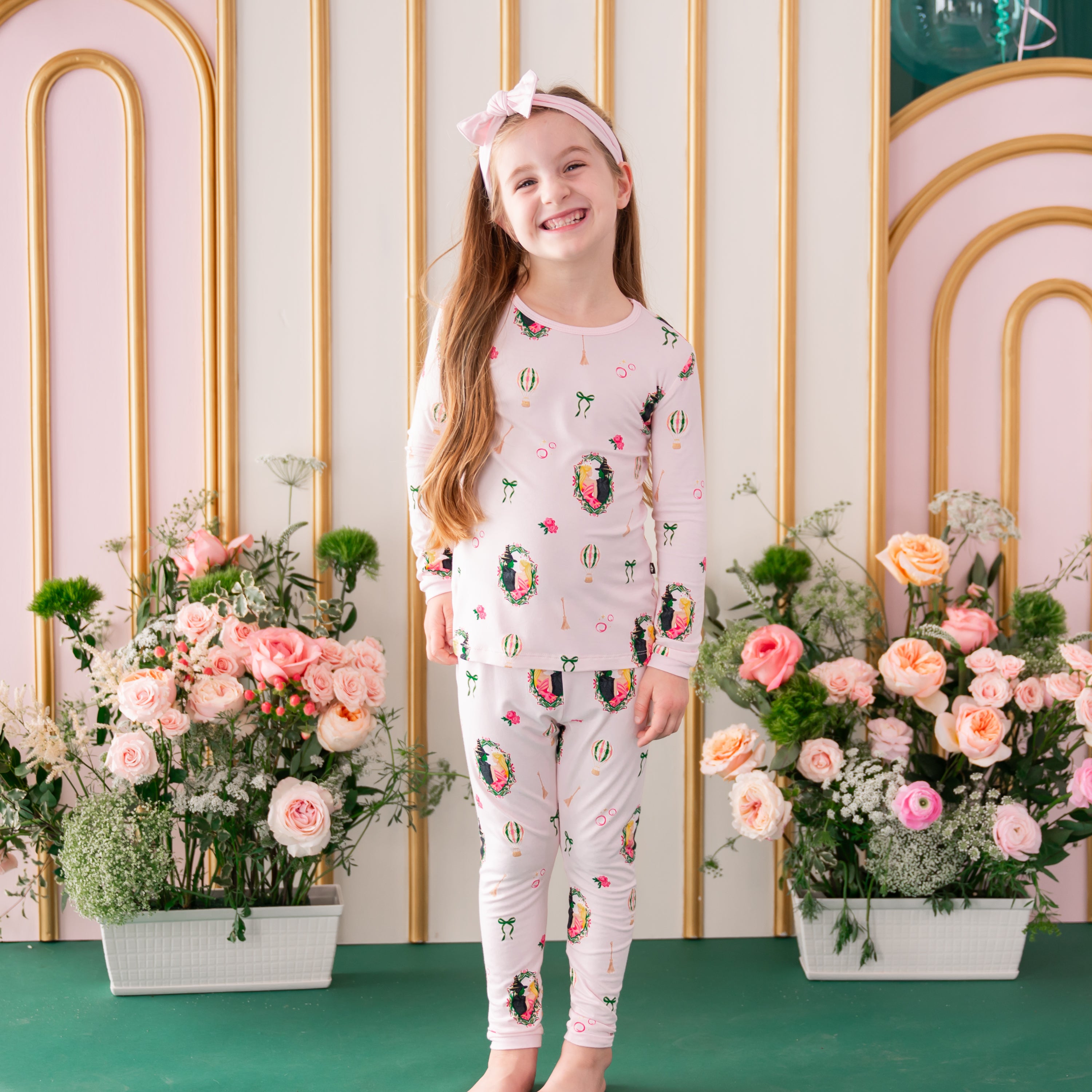 Young smiling girl wearing the breathable Long Sleeve Pajamas in Wicked from Kyte Baby standing in front of a floral backdrop