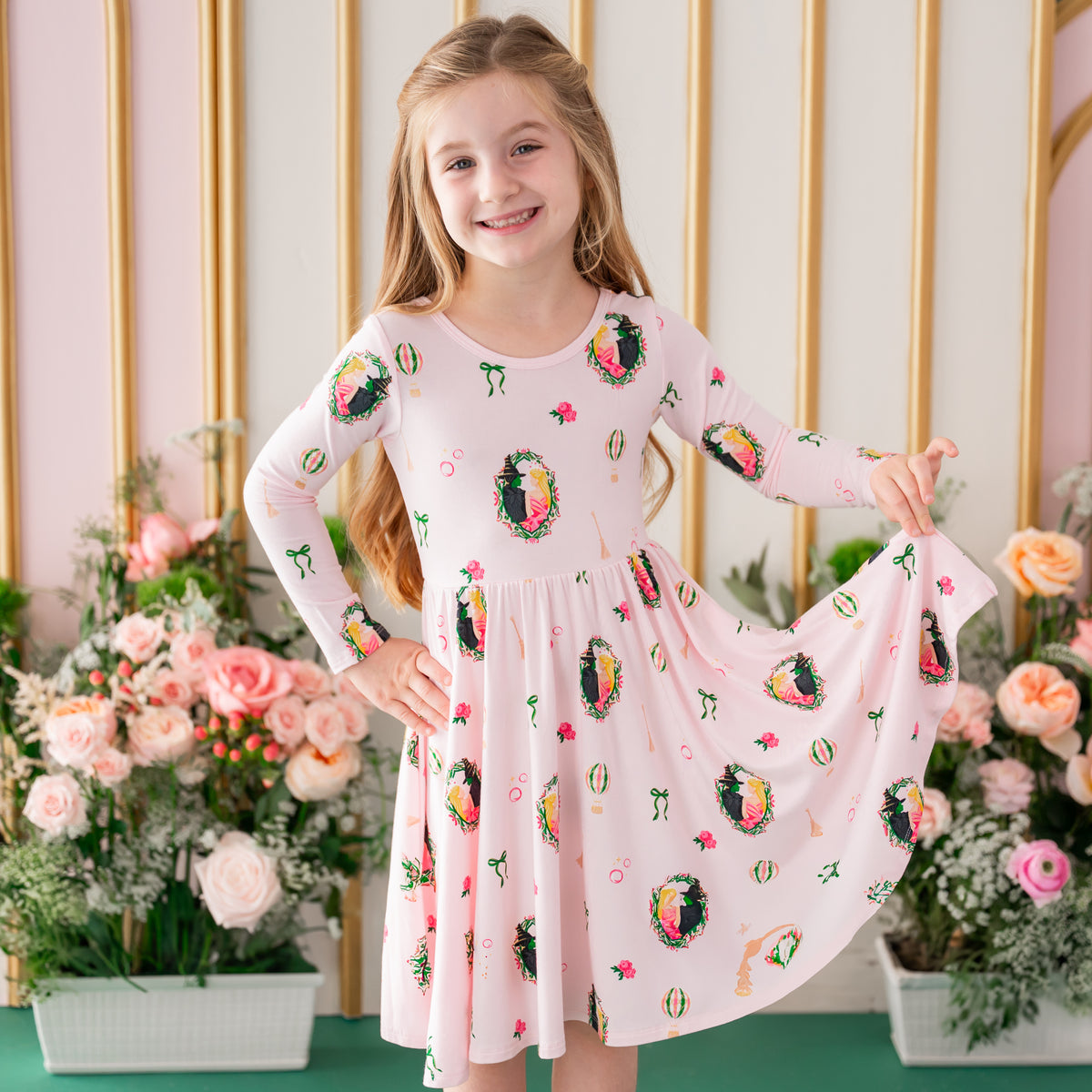 Young girl wearing the Long Sleeve Twirl Dress in Wicked holding the hem up standing in front of a floral backdrop