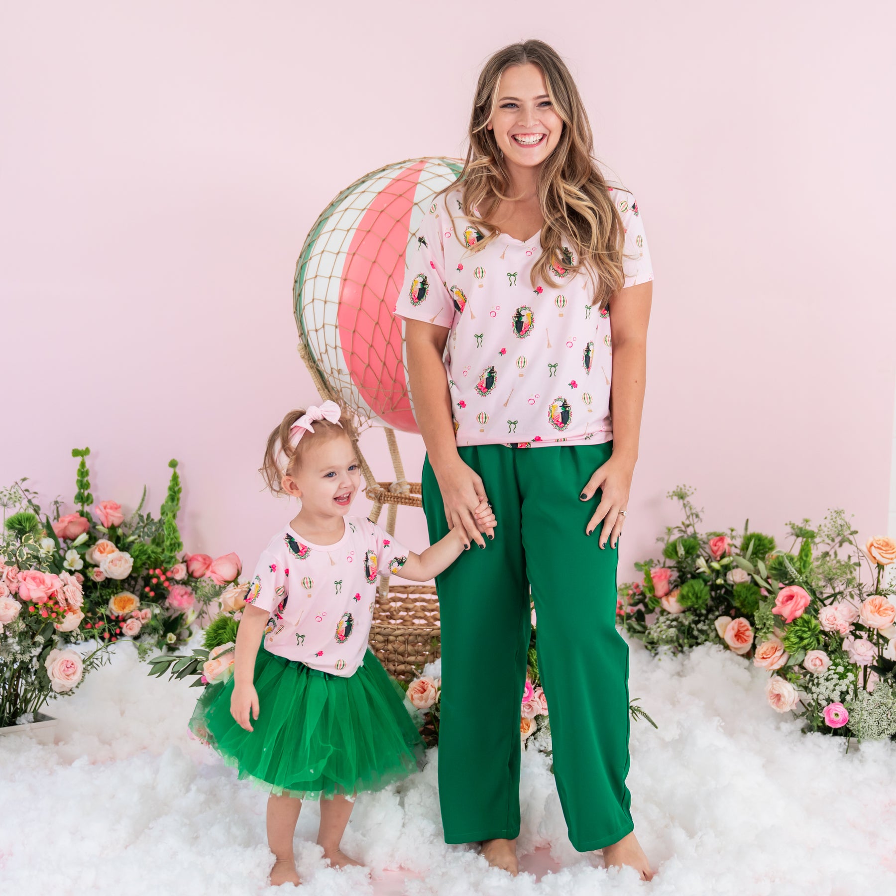 Young girl wearing the Toddler Basic Tee in Wicked and green tulle skirt holding her mothers hand who is wearing a matching women' V-neck and green pants both standing in front of a prop hot air balloon and flower arrangements