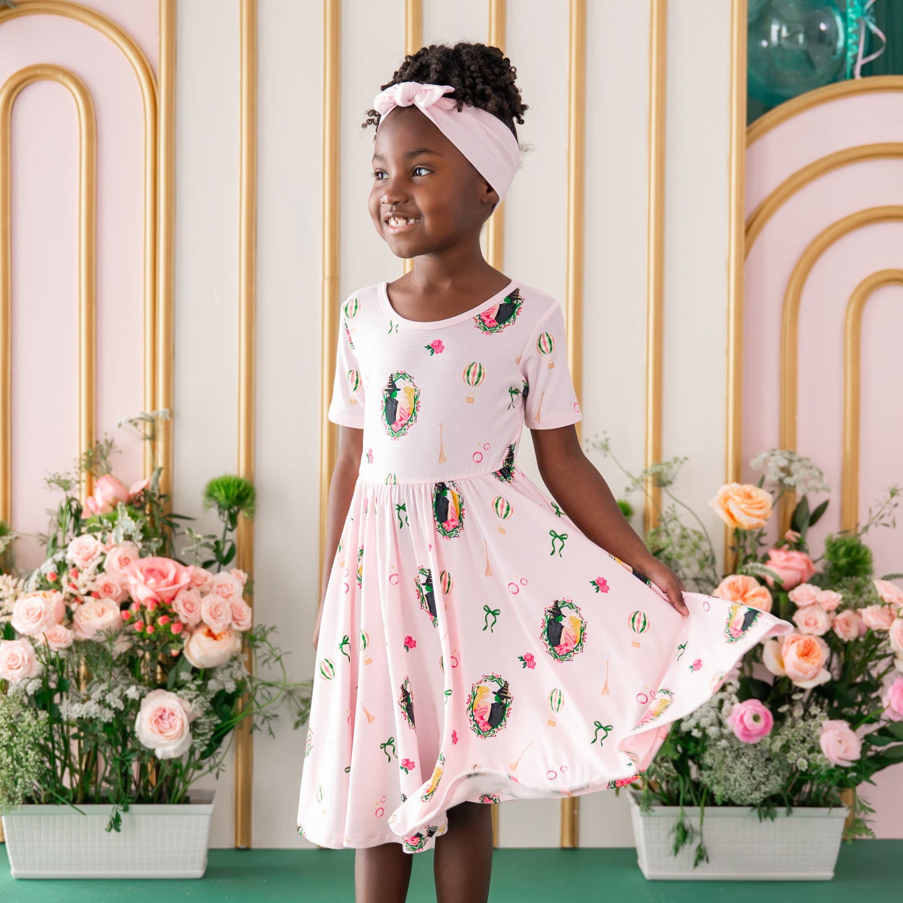 Smiling girl wearing the Twirl Dress in Wicked paired with a light pink bow standing in front of a floral backdrop
