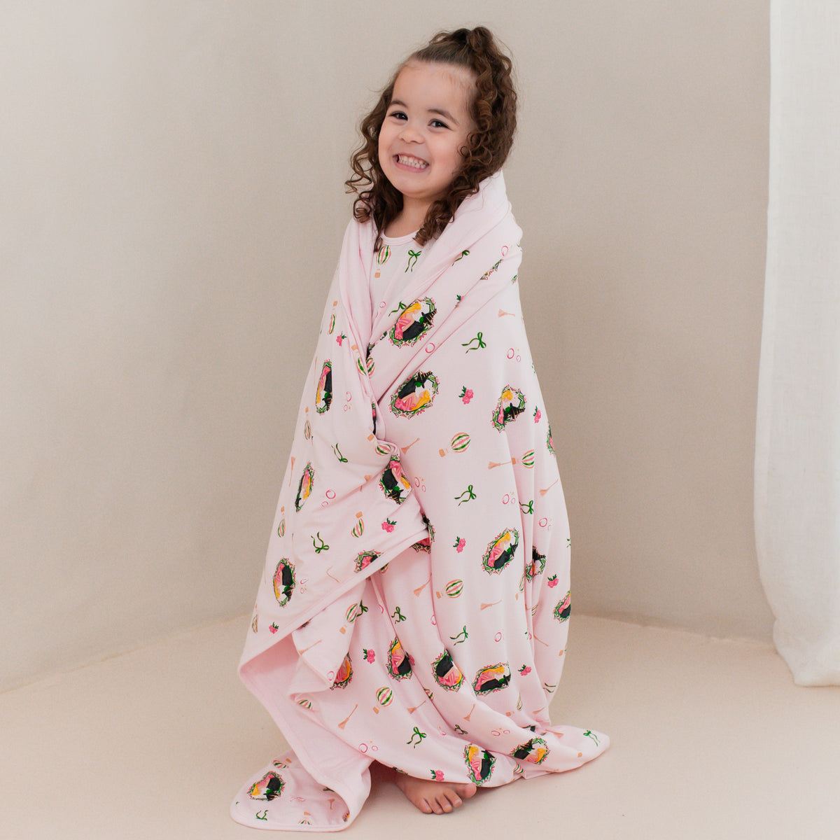 Young girl standing in front of a beige wall with the soft and breathable Toddler Blanket in Wicked 1.0 from Kyte Baby wrapped around her